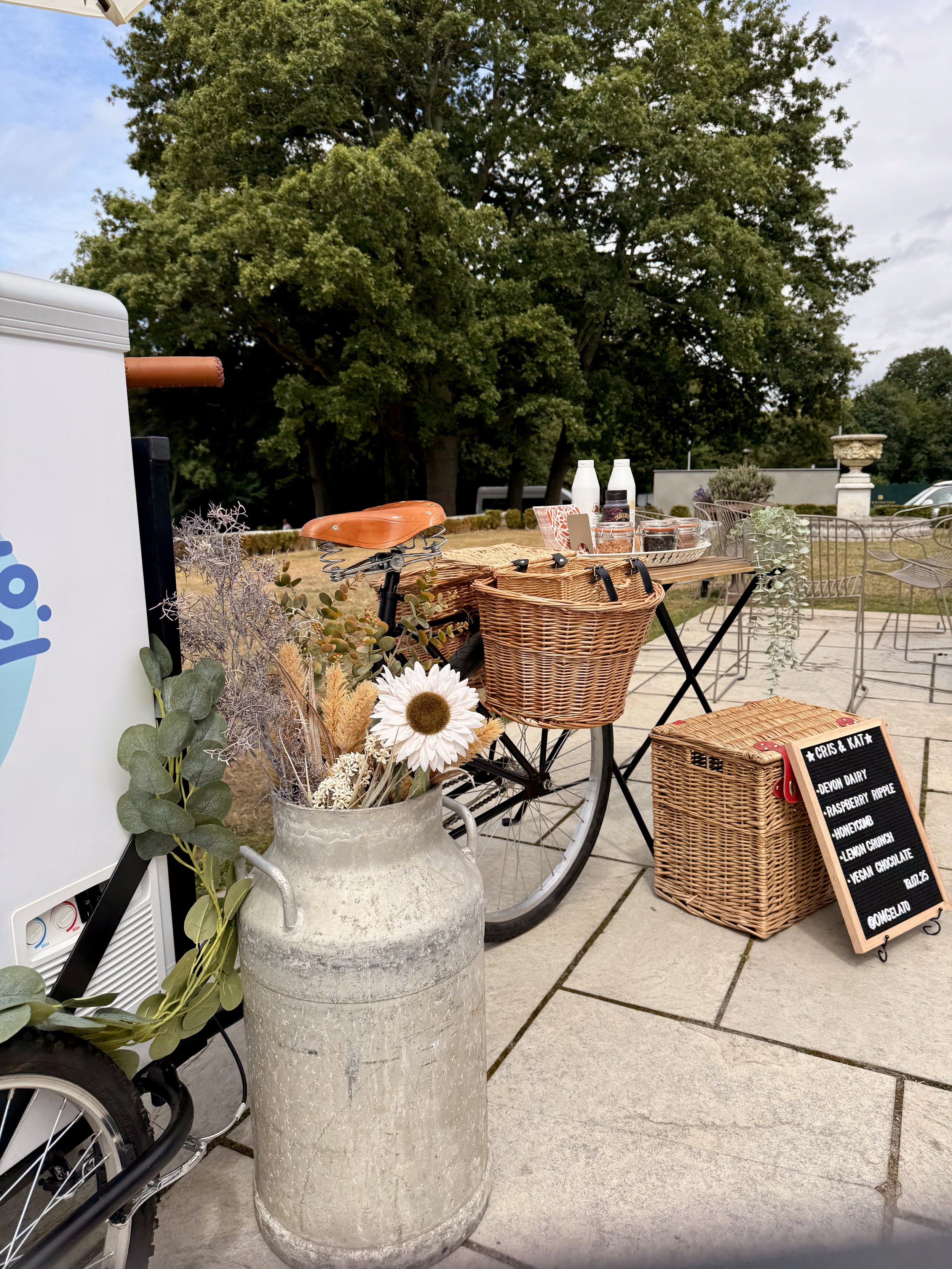 A vintage bicycle with a wicker basket on the front, parked on a patio next to a large ceramic vase filled with dried flowers, in front of an outdoor ice cream stand with a chalkboard menu and various toppings. Trees and outdoor furniture are visible
