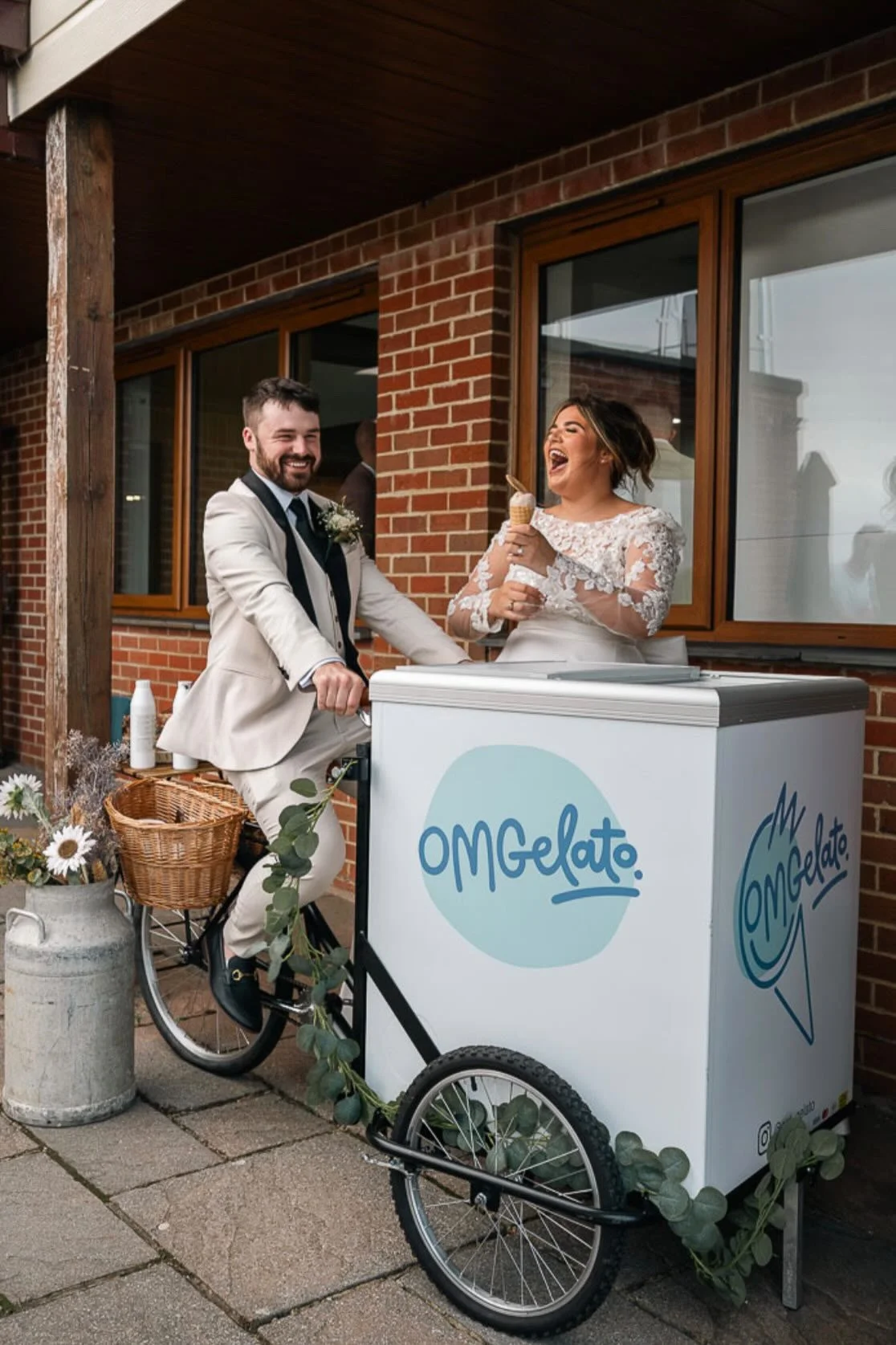 A couple dressed in wedding attire enjoying ice cream from a gelato cart outside a brick building.