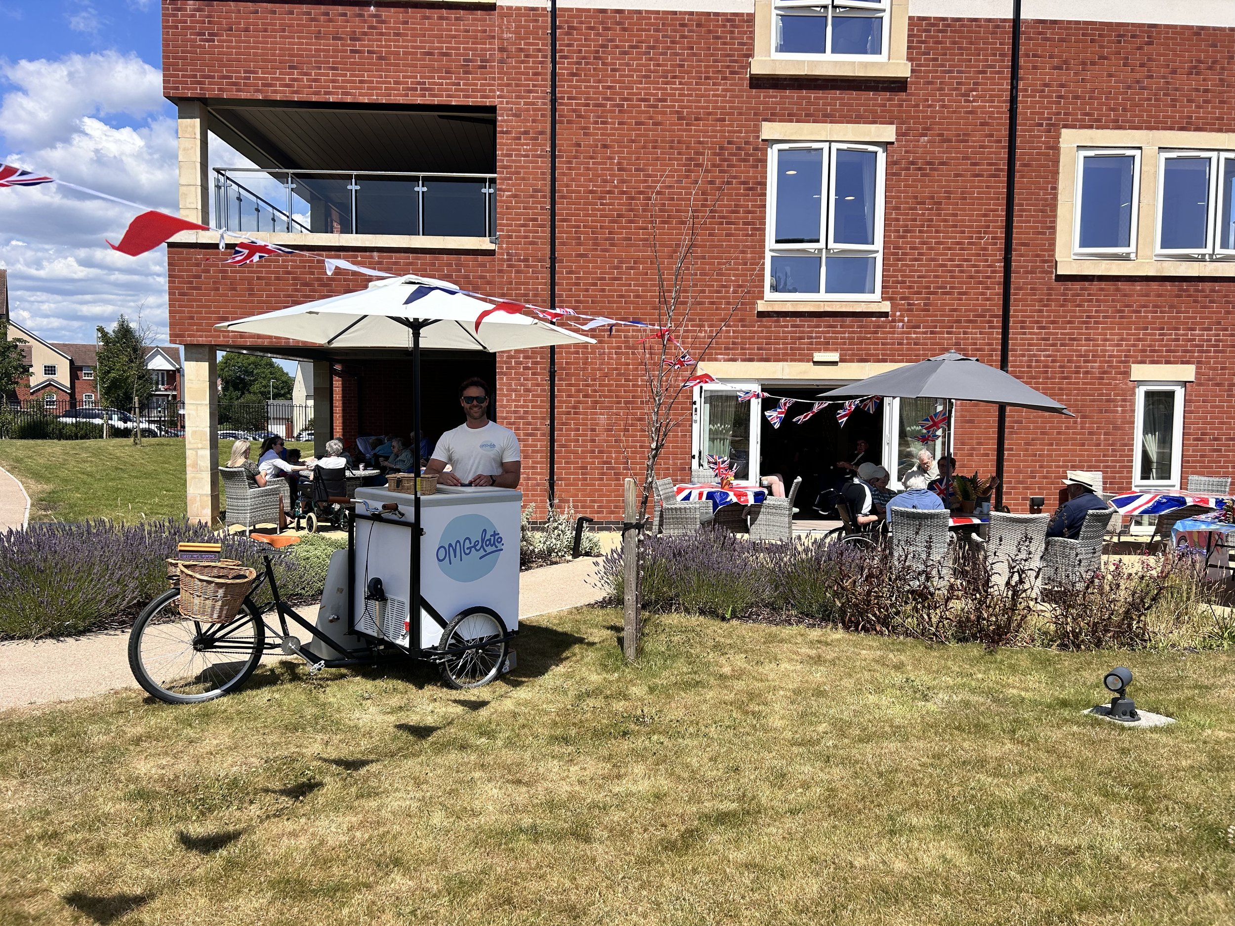 People enjoying an outdoor gathering with umbrellas and British flags on a patio in front of a red brick building, with an ice cream bike in the foreground.