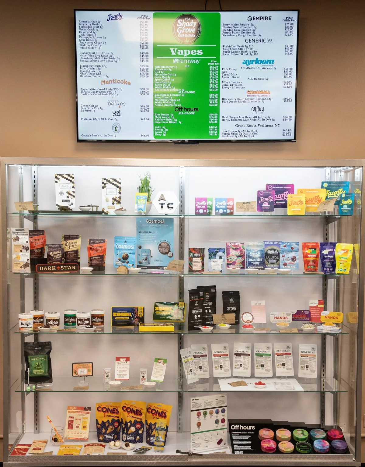 Display case with various cannabis products including edibles, tinctures, and packages, and an electronic menu screen above showing cannabis strains and prices.