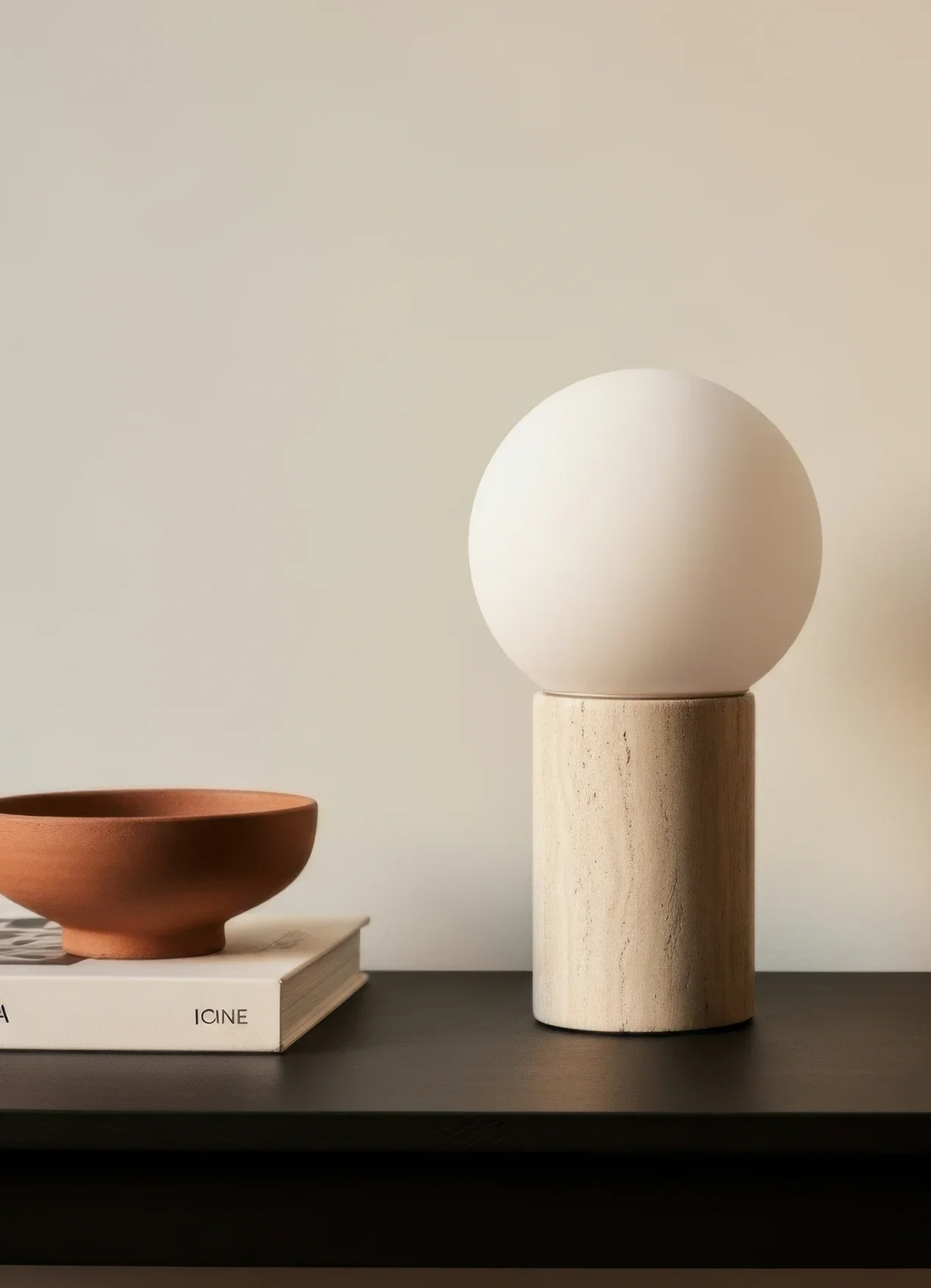A minimalist display with a terracotta-colored bowl, a book, and a modern lamp with a spherical white shade on a wooden base, set on a black table against a neutral wall.