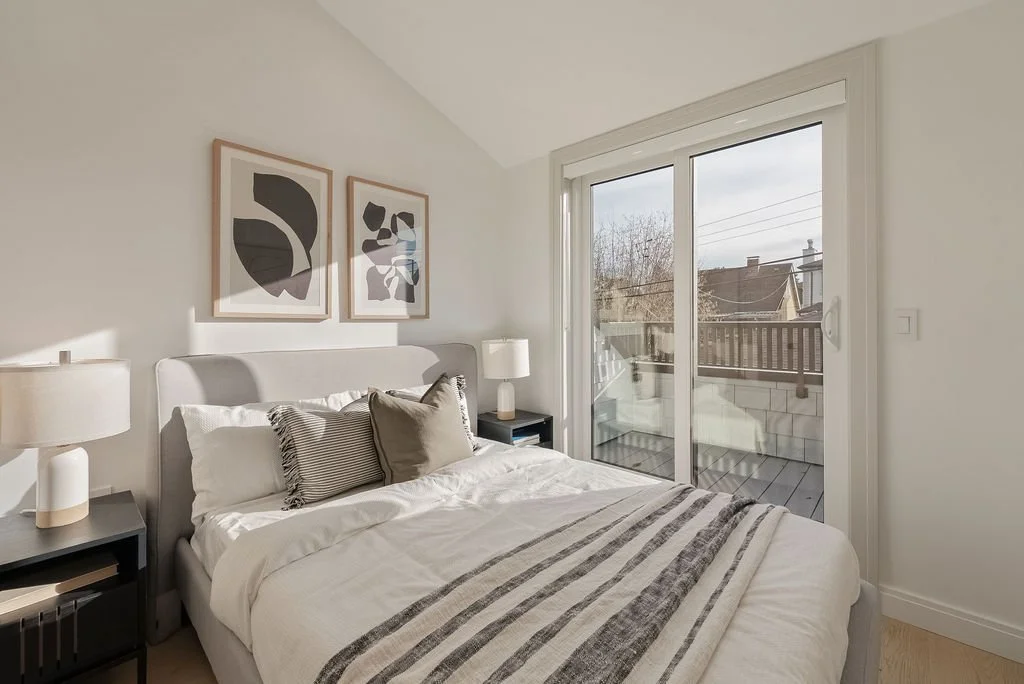 Bright bedroom with a bed, nightstands, and lamps, near a sliding glass door leading to a small balcony outside with some buildings in view.