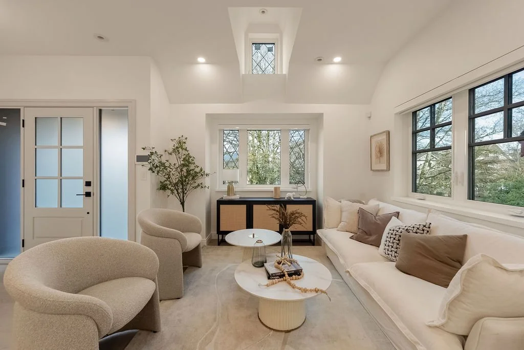 Bright living room with white walls, large windows, beige sofa with pillows, two armchairs, a small round coffee table, and a sideboard with plants and decorative items.