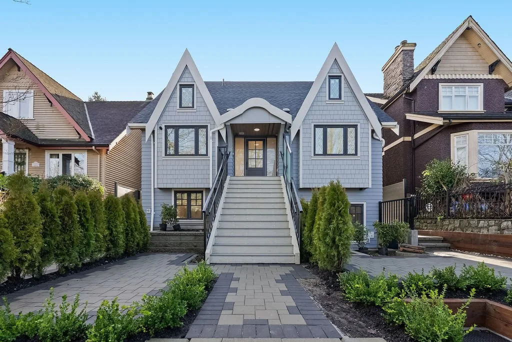 A modern two-story house with a steep gable roof, gray siding, and stairs leading up to the front door. It is flanked by green shrubs and landscaping. The house is situated between two other houses with different architectural styles.