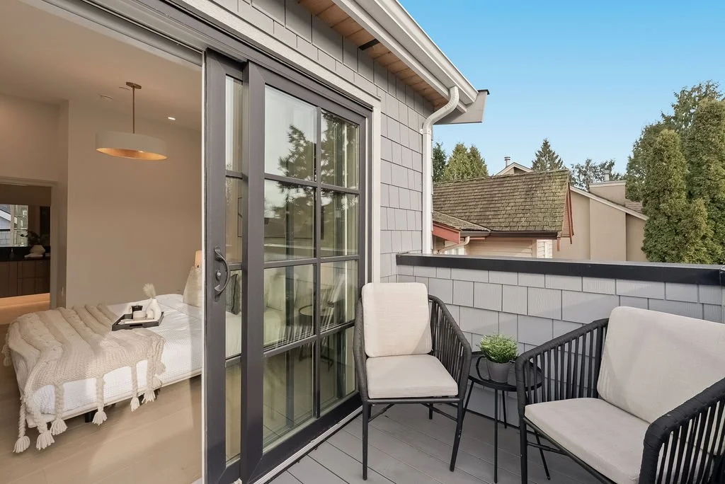 A small outdoor balcony with two white cushioned chairs around a small table with a potted plant. The balcony is attached to a house with a sliding glass door leading inside, and the house has gray siding. The view includes neighboring houses and trees.