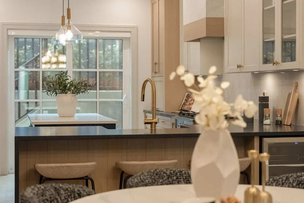 Modern kitchen with white cabinets, black countertop, gold faucet, and a potted plant on the countertop. Pendant lights hang above a small dining table with a plant, and a sliding glass door leads outside.