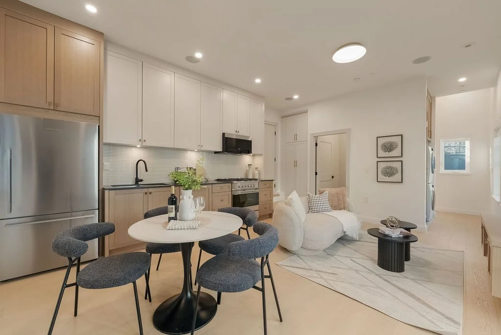 Open-concept kitchen and living area with light wood and white cabinetry, stainless steel refrigerator, black countertop appliances, a dining table with five dark gray chairs, and a cozy seating area with a white curved sofa, small black coffee table, and wall art.