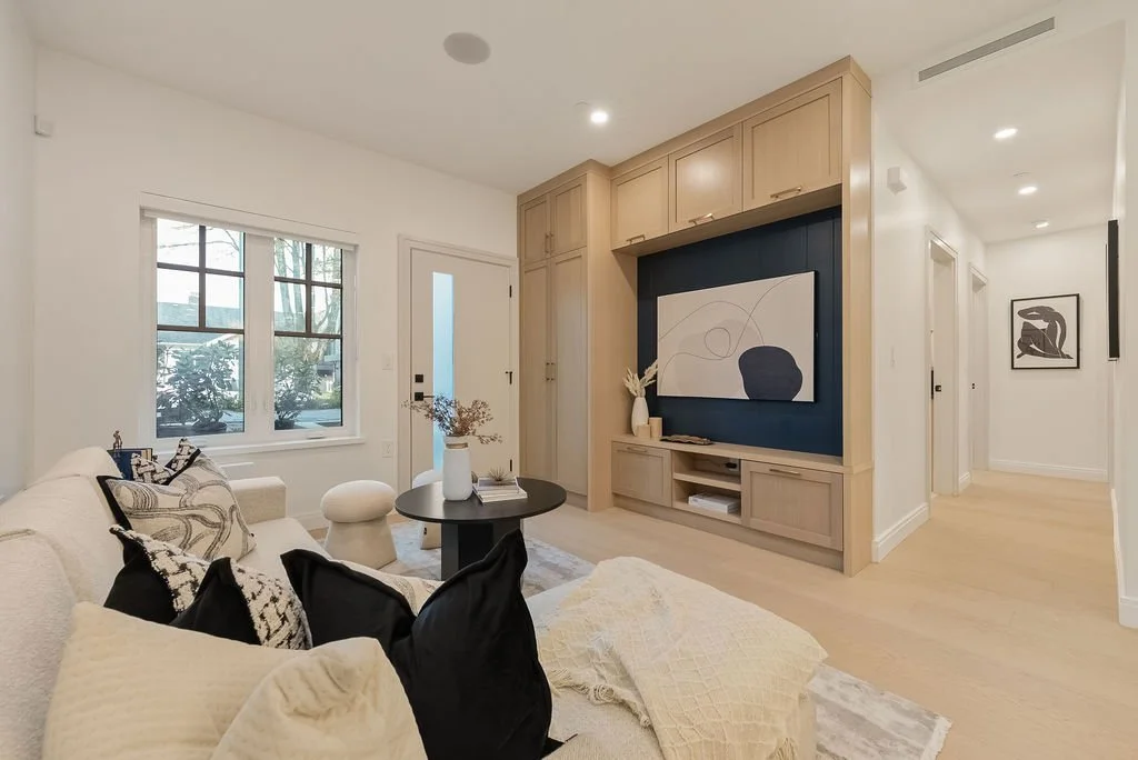 Living room with cream-colored sofa, decorative pillows, round black coffee table with books and a vase, window with view of trees, wooden built-in cabinets with decorative art, and hallway with framed artwork.