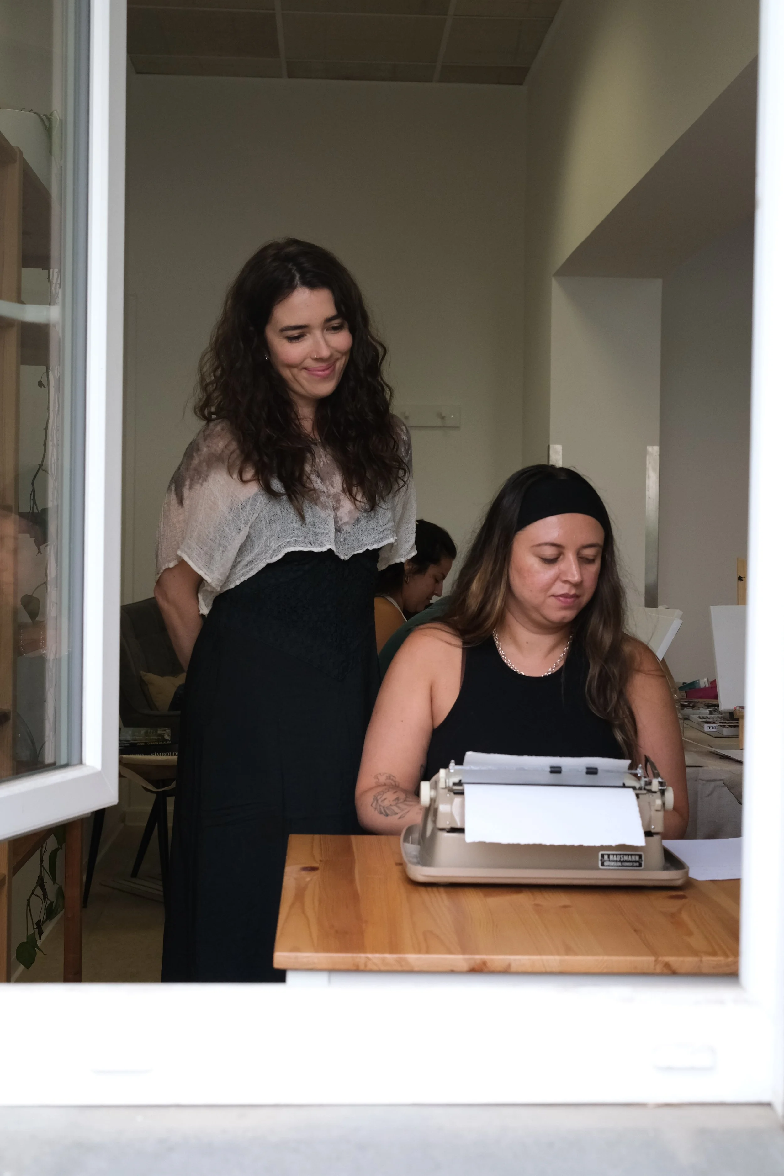 Two women at a wooden table using a typewriter, seen through a window with two women in the background, one standing and one sitting, in a room with beige walls and a ceiling with brown tiles.