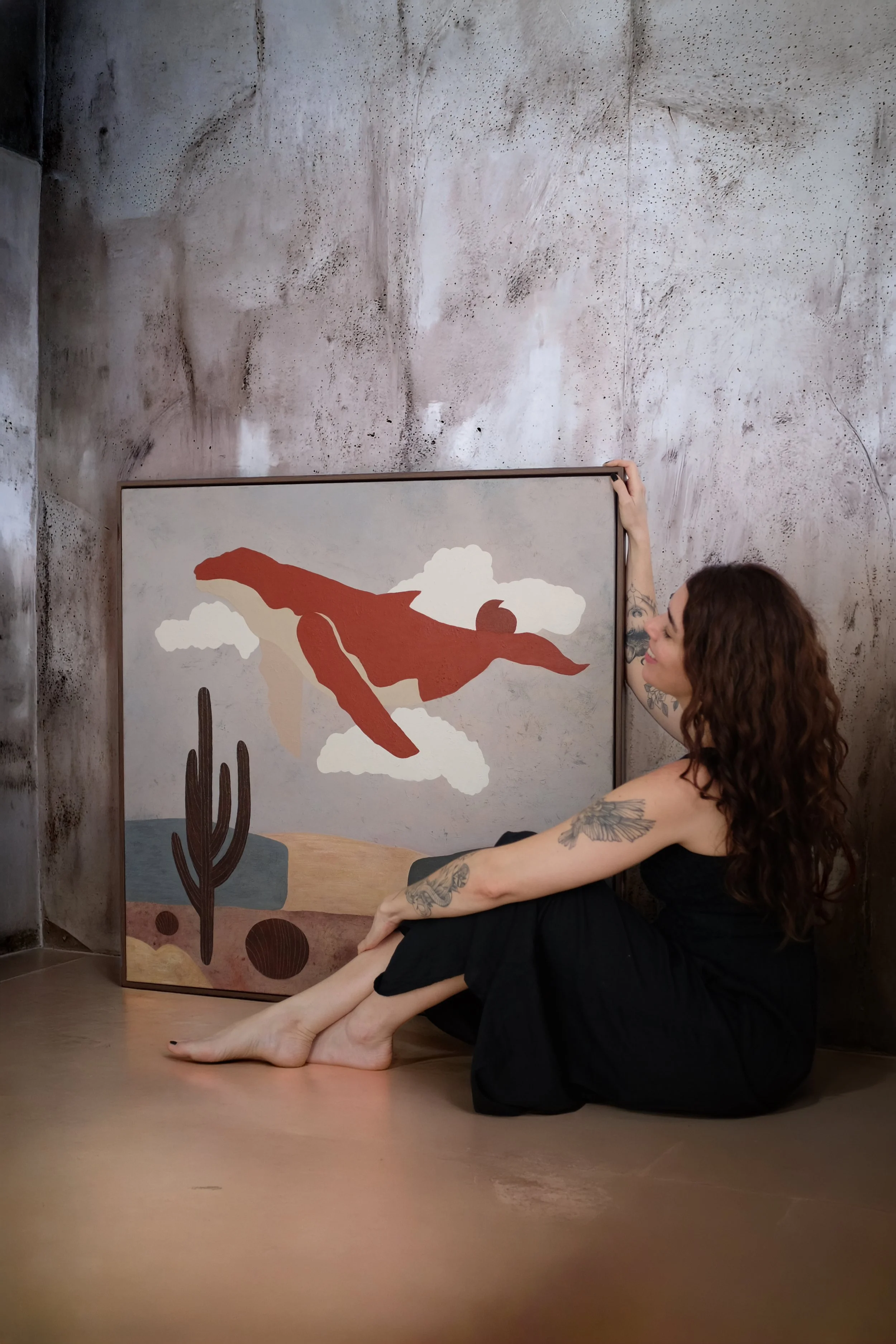 A woman with curly brown hair, tattoos on her arms, and wearing a black dress sitting on the floor, holding a framed piece of artwork featuring a stylized desert scene with a red bird and cacti, against a textured concrete wall.