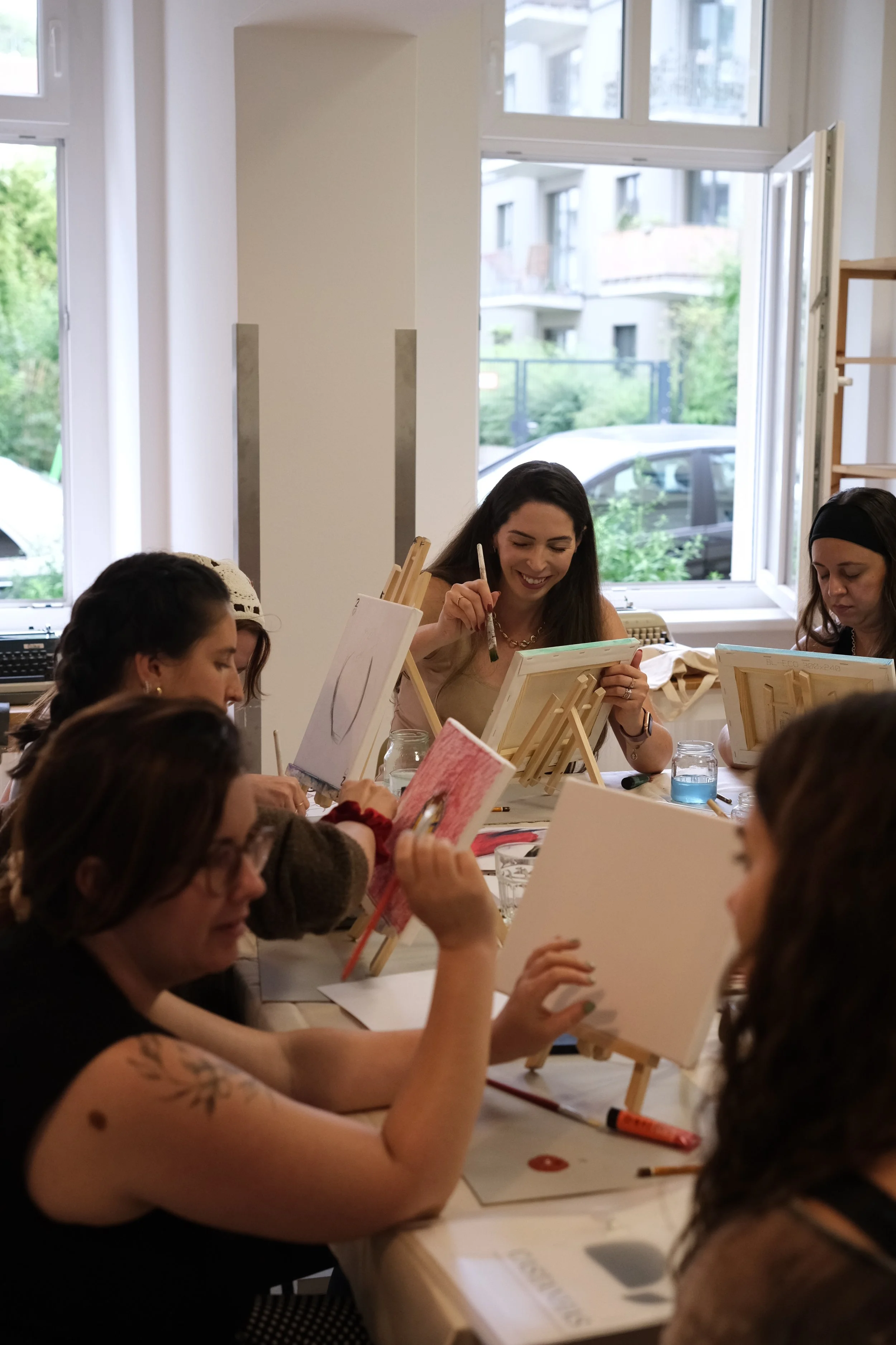 A group of women sitting around a table, painting on canvases supported on small easels in a well-lit room with large windows.