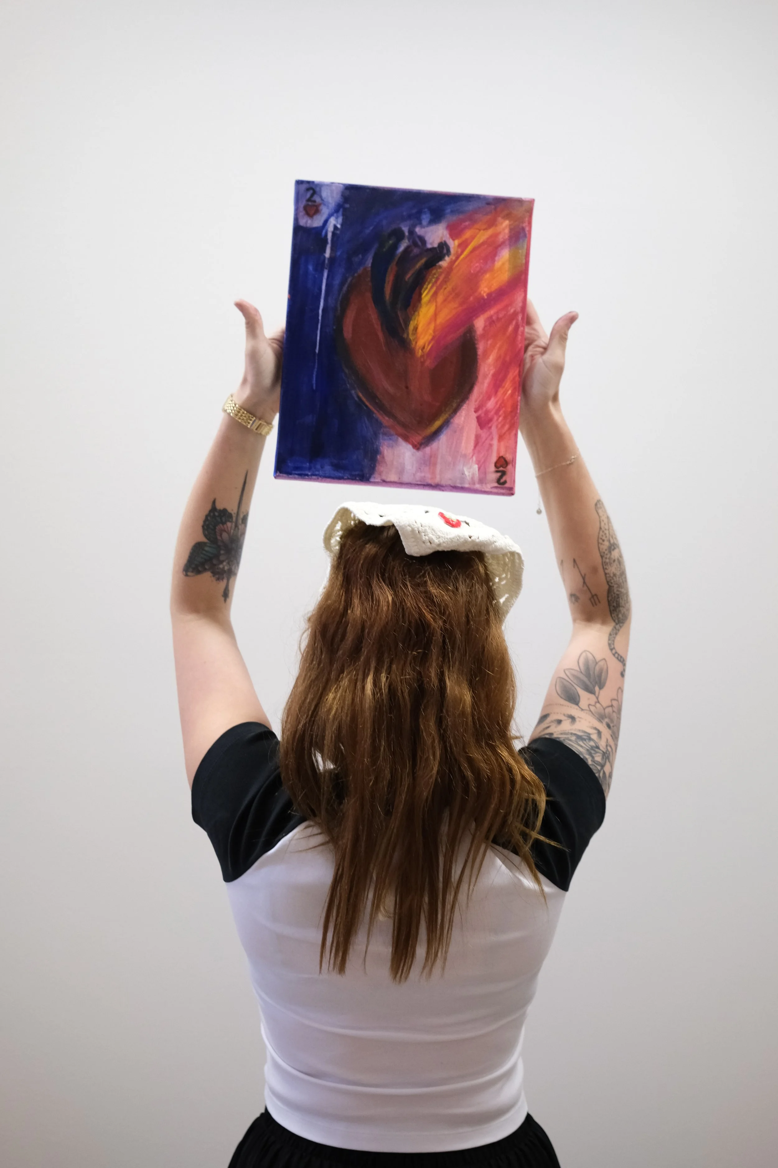 Person holding a colorful abstract painting above their head, with tattoos on arms, wearing a hat and a short-sleeved shirt, standing against a plain white wall.