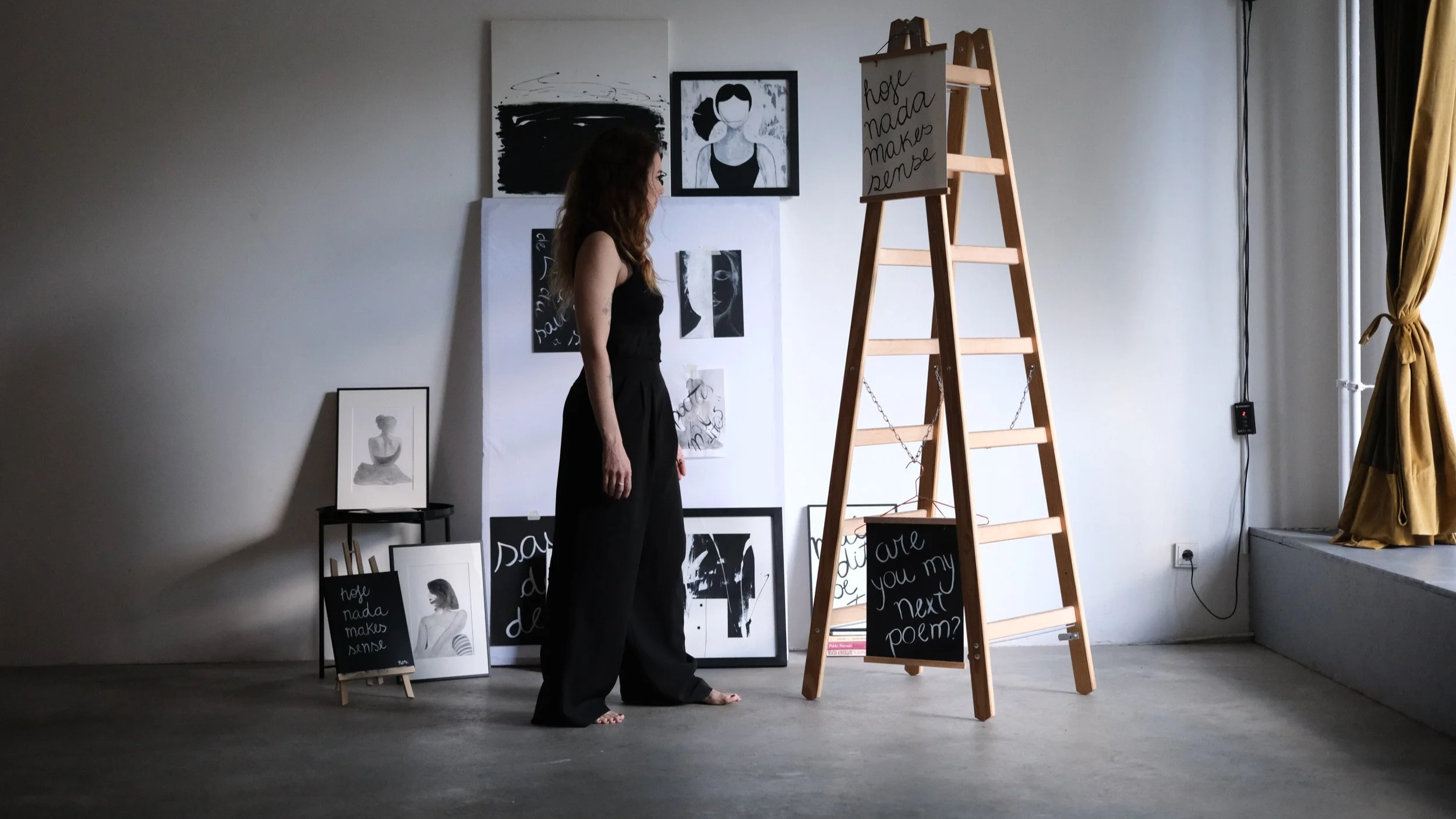 A woman standing barefoot in an art studio with black and white artwork and signs, including one on a wooden ladder that says, "Hoy nada makes sense" and another hanging that says, "Are you my next poem?"