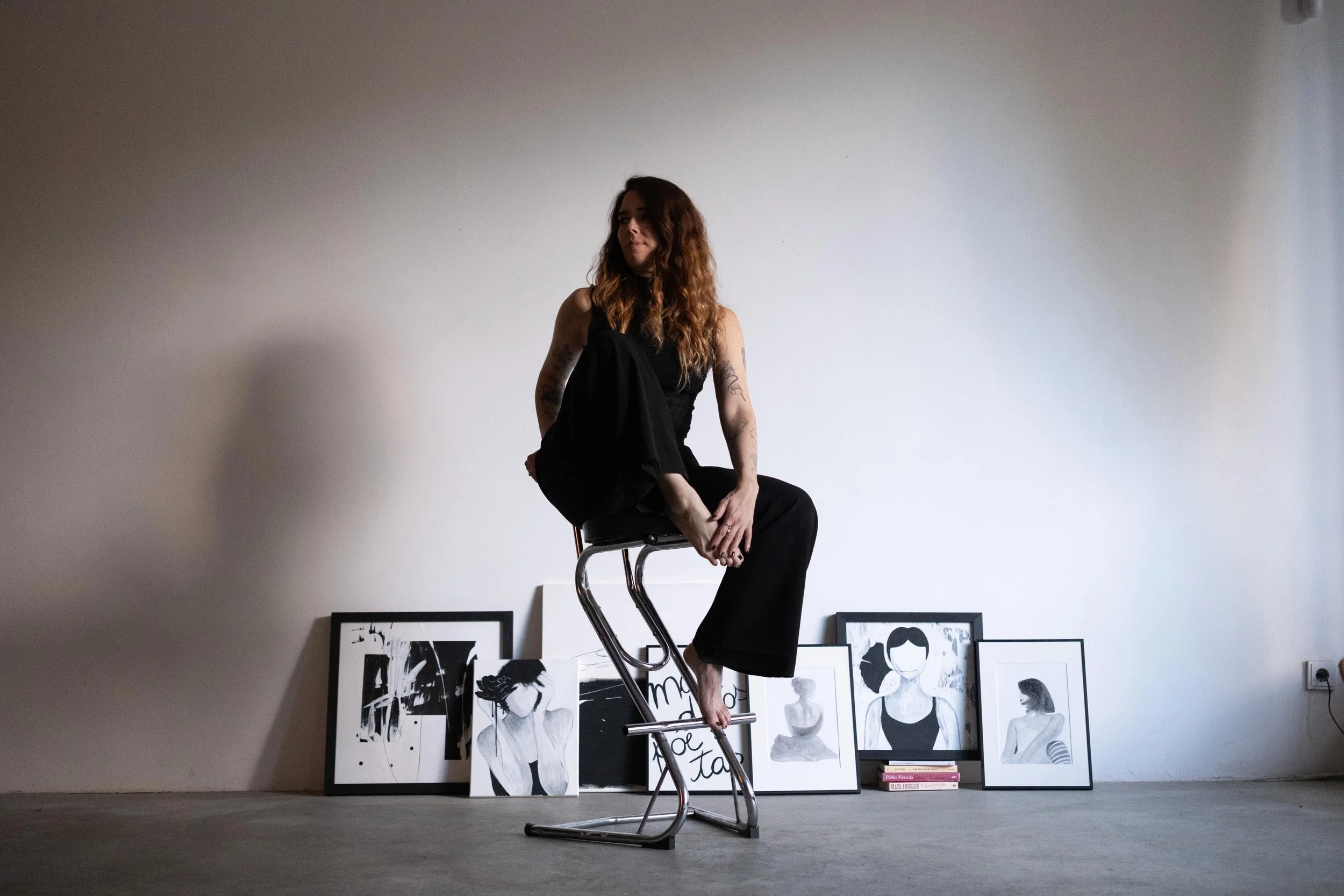 A woman with long, wavy hair and tattoos, dressed in black, sitting on a modern metal chair in an art studio with framed black-and-white artwork on the floor against a plain wall.