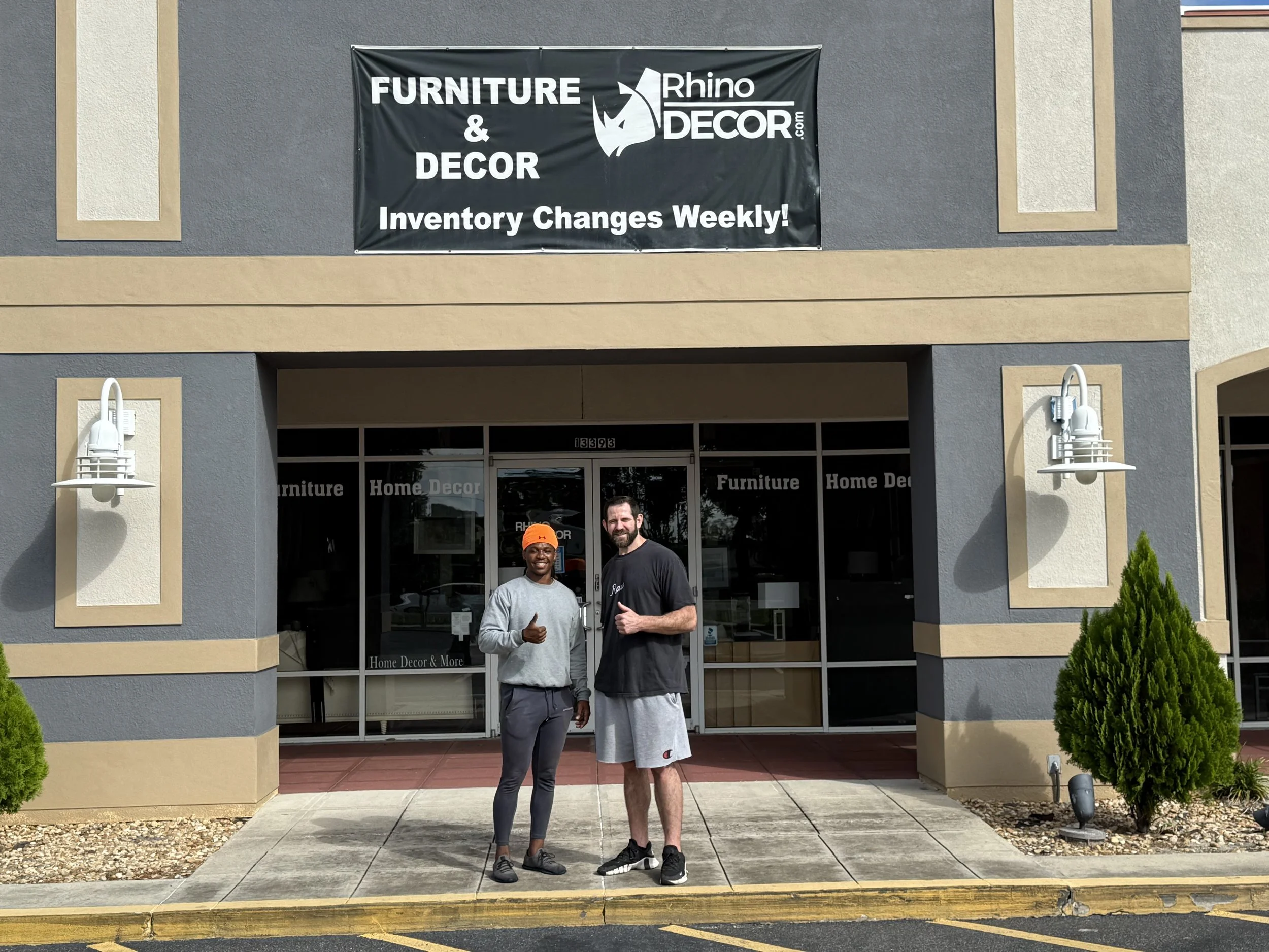 Two men standing outside a furniture and home decor store posing for a photo, one giving a thumbs-up, storefront sign above.