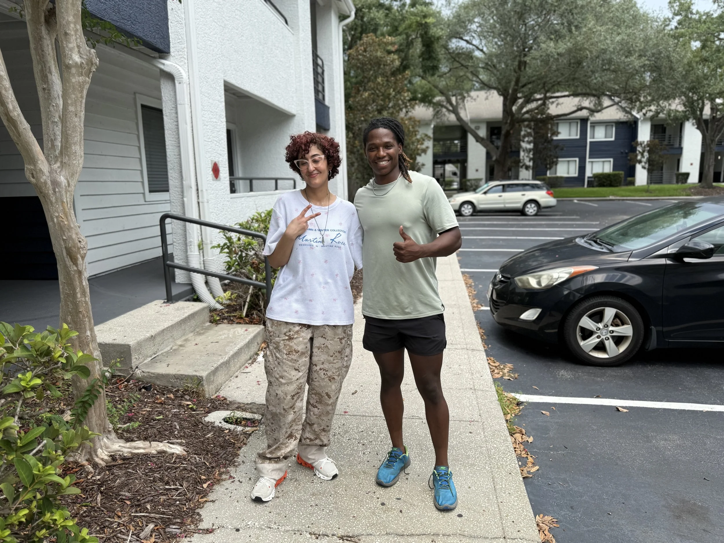 Two people standing outside on a sidewalk near parked cars, smiling and making gestures, with apartment buildings and trees in the background.