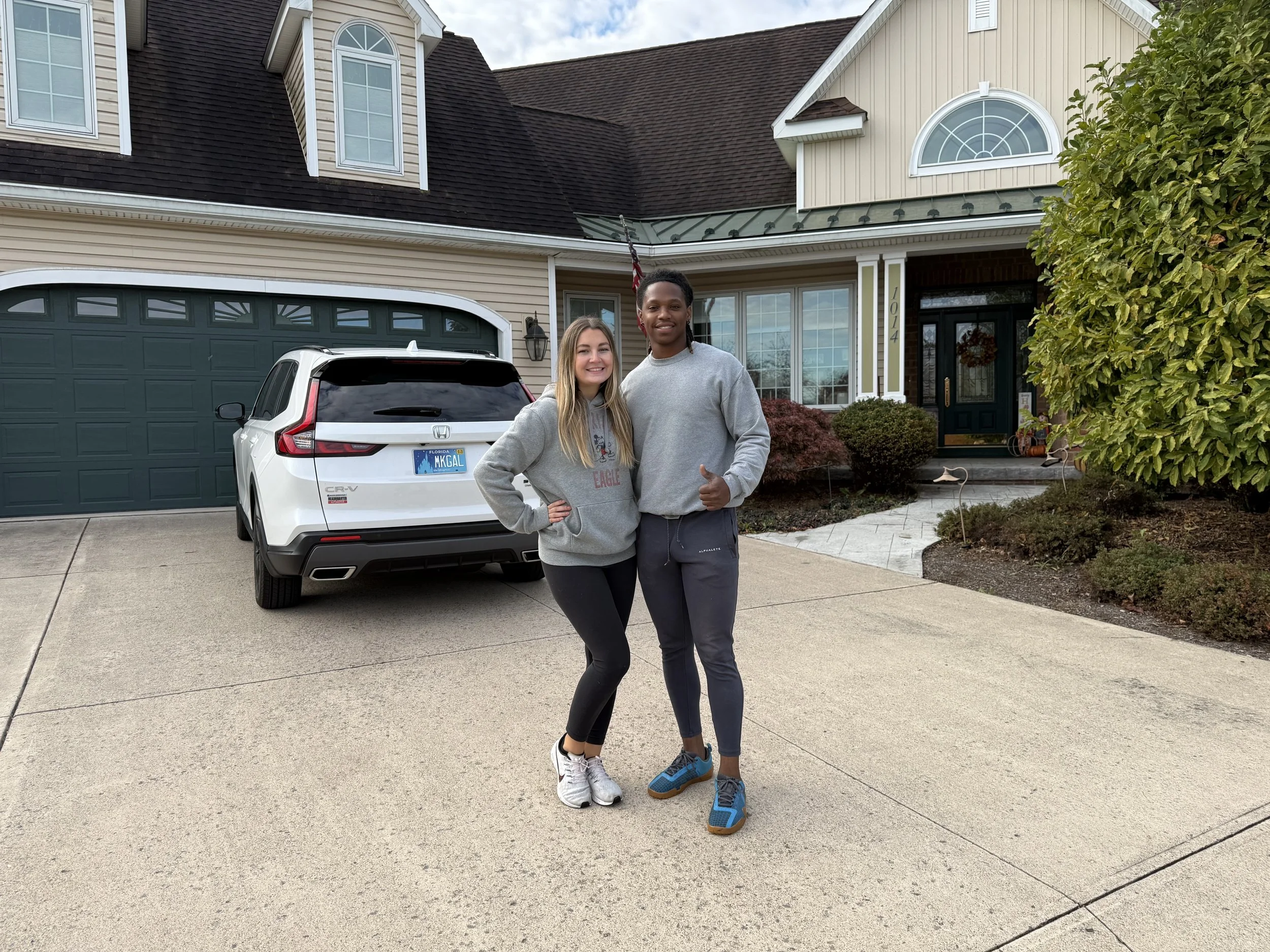 A young woman and man standing in front of a white Honda SUV parked in a driveway, in front of a house with a green front door, window, and tan siding, with landscaping including bushes and a tree.