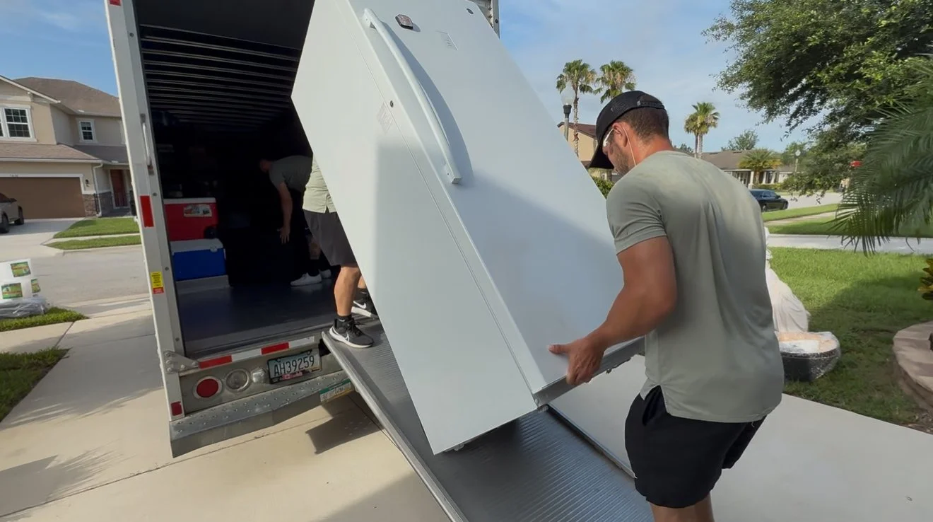 Two men unloading a refrigerator from a moving truck onto a driveway in a suburban neighborhood.