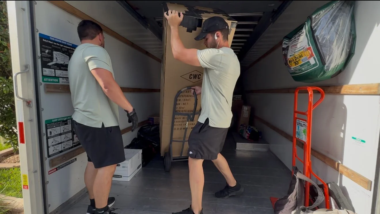 Two men are loading a moving box into a moving truck. One man is holding the box and the other is helping to lift it. Inside the truck, there is a green bag on the right side and a red hand truck on the right.