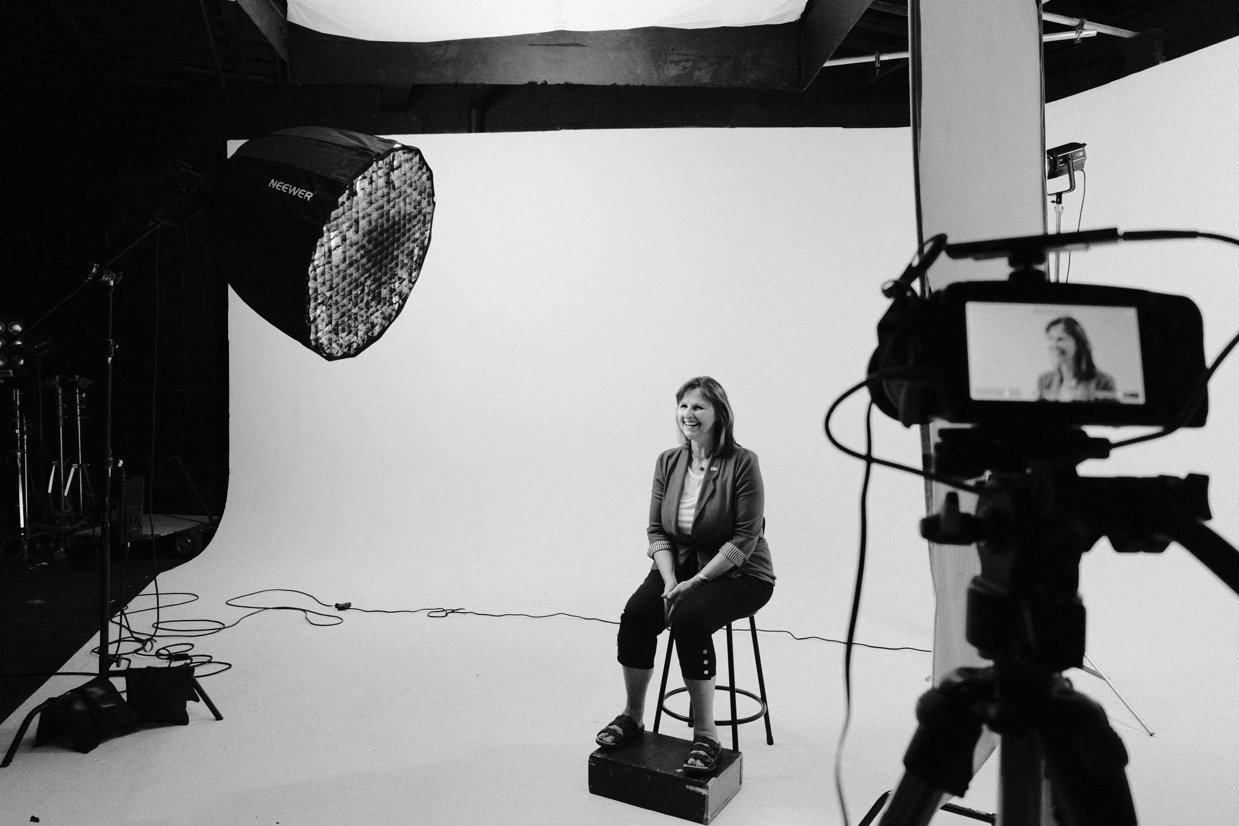 Black and white photo of a woman sitting on a stool in front of a plain white backdrop in a photography studio, with studio lights and camera equipment around her.