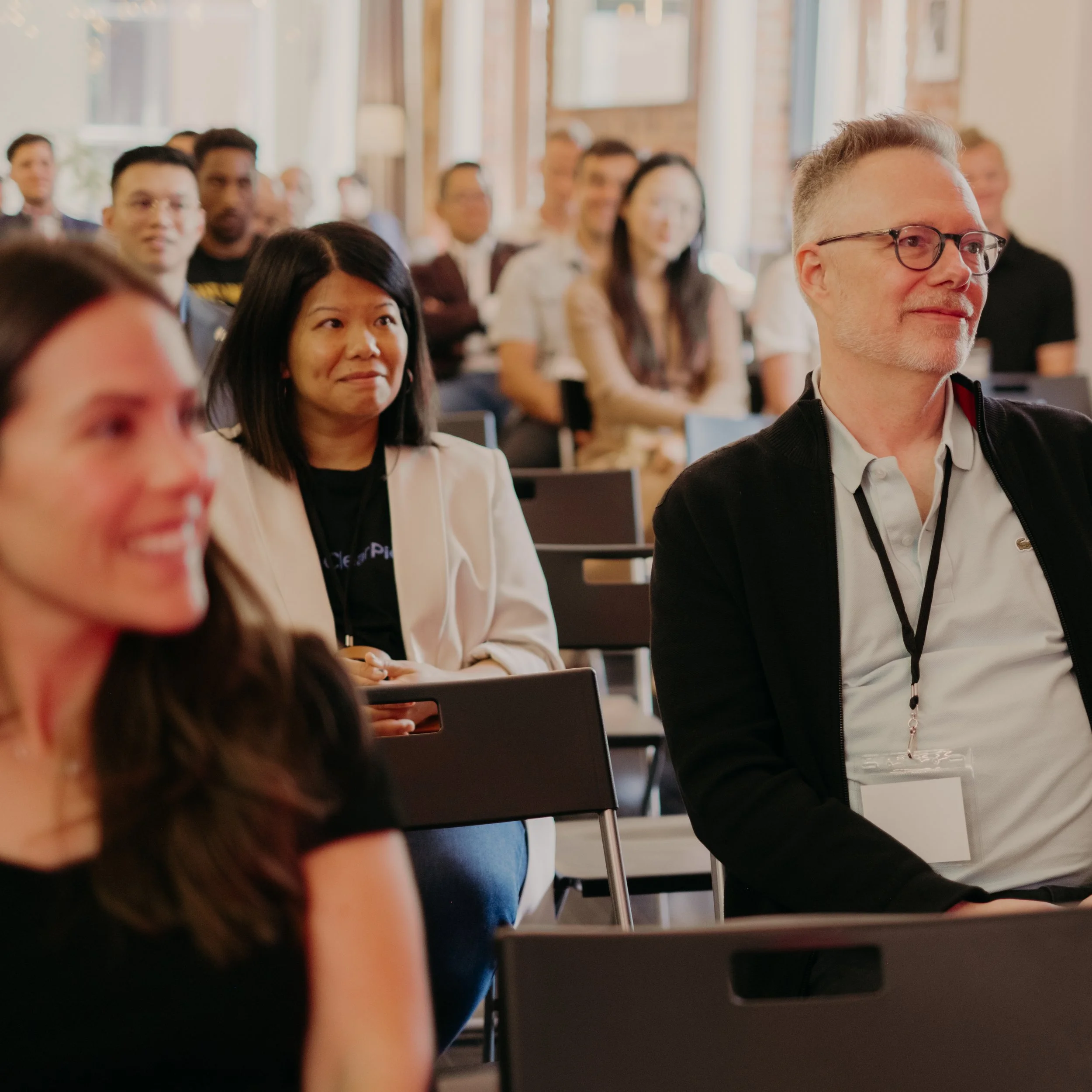 People attending a conference or seminar in a bright, modern room, sitting on black chairs and listening attentively.