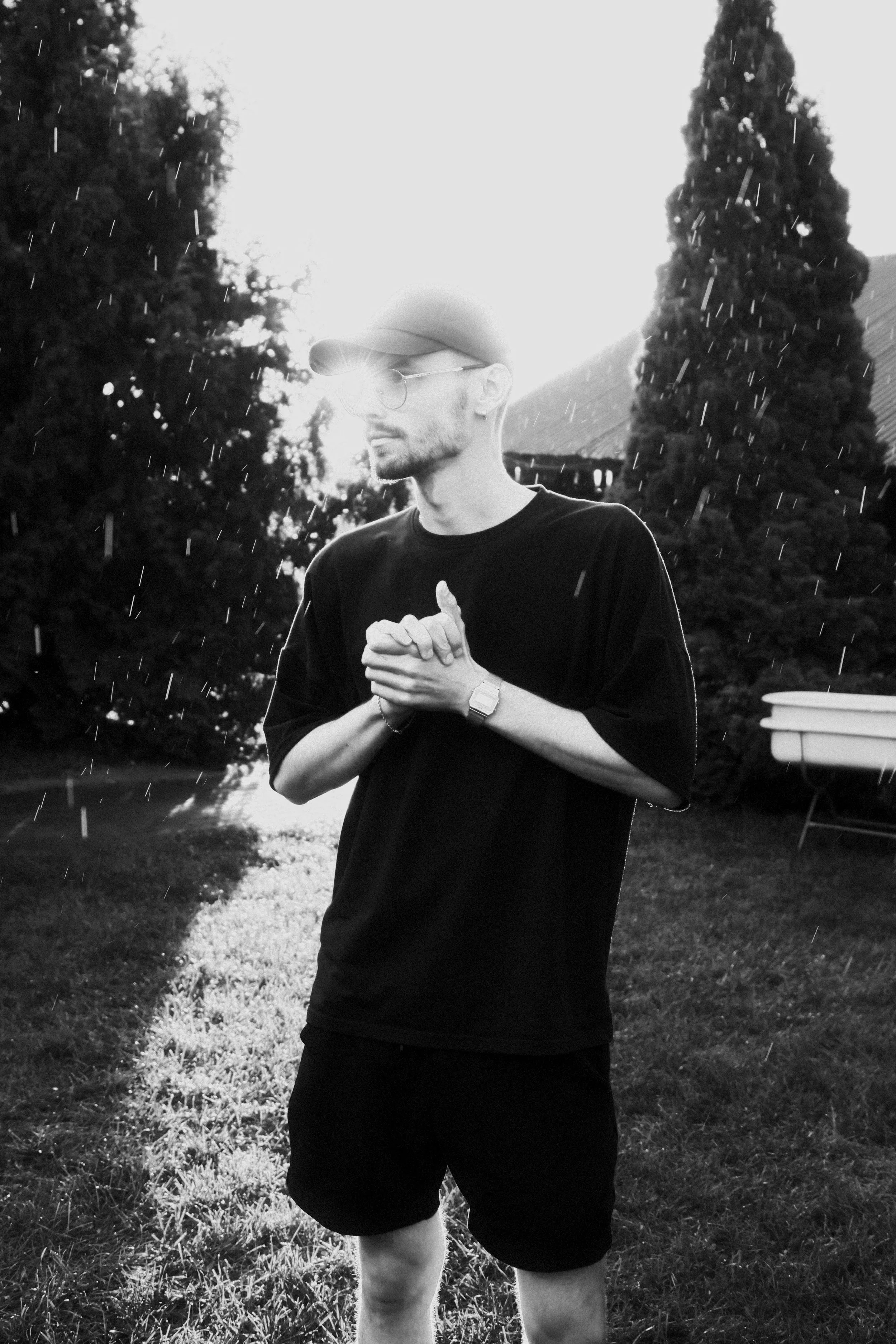 A man standing outside on a grassy area during rain, wearing a baseball cap, sunglasses, and a T-shirt, with trees and a building in the background.