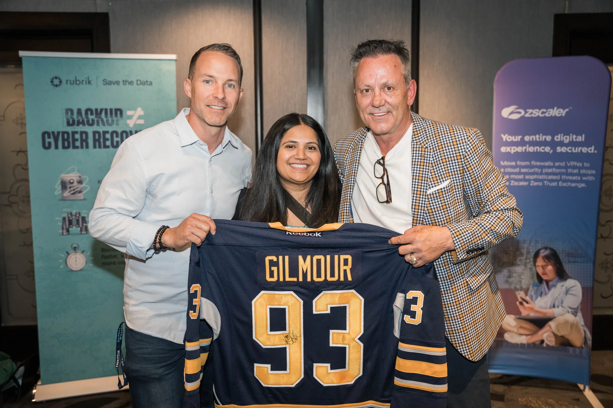 Three people smiling and holding a hockey jersey with the name Gilmour and the number 93, standing in front of promotional banners at an event.