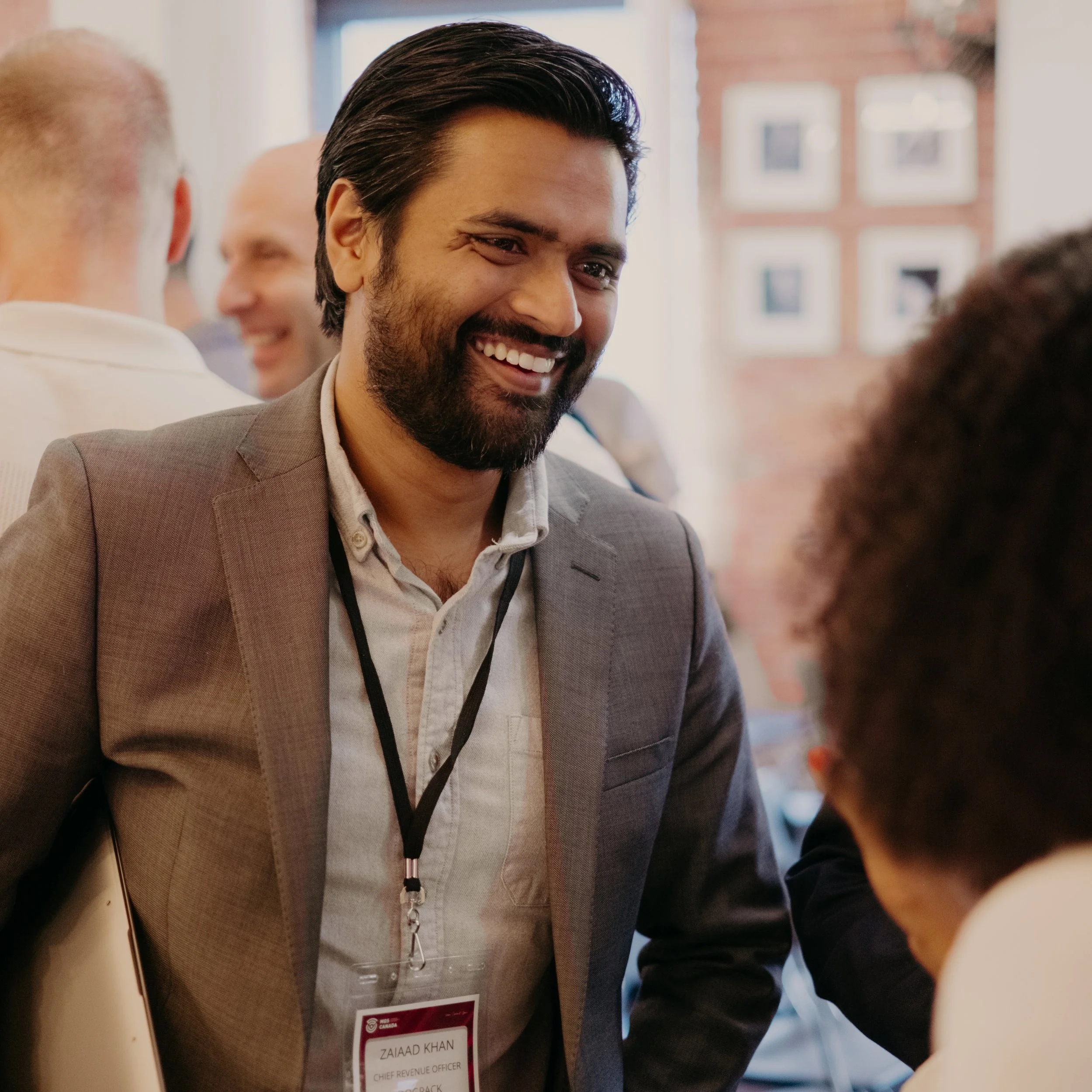 Man with dark hair, beard, wearing a gray blazer and lanyard with ID badge, smiling and talking to someone at a professional event.