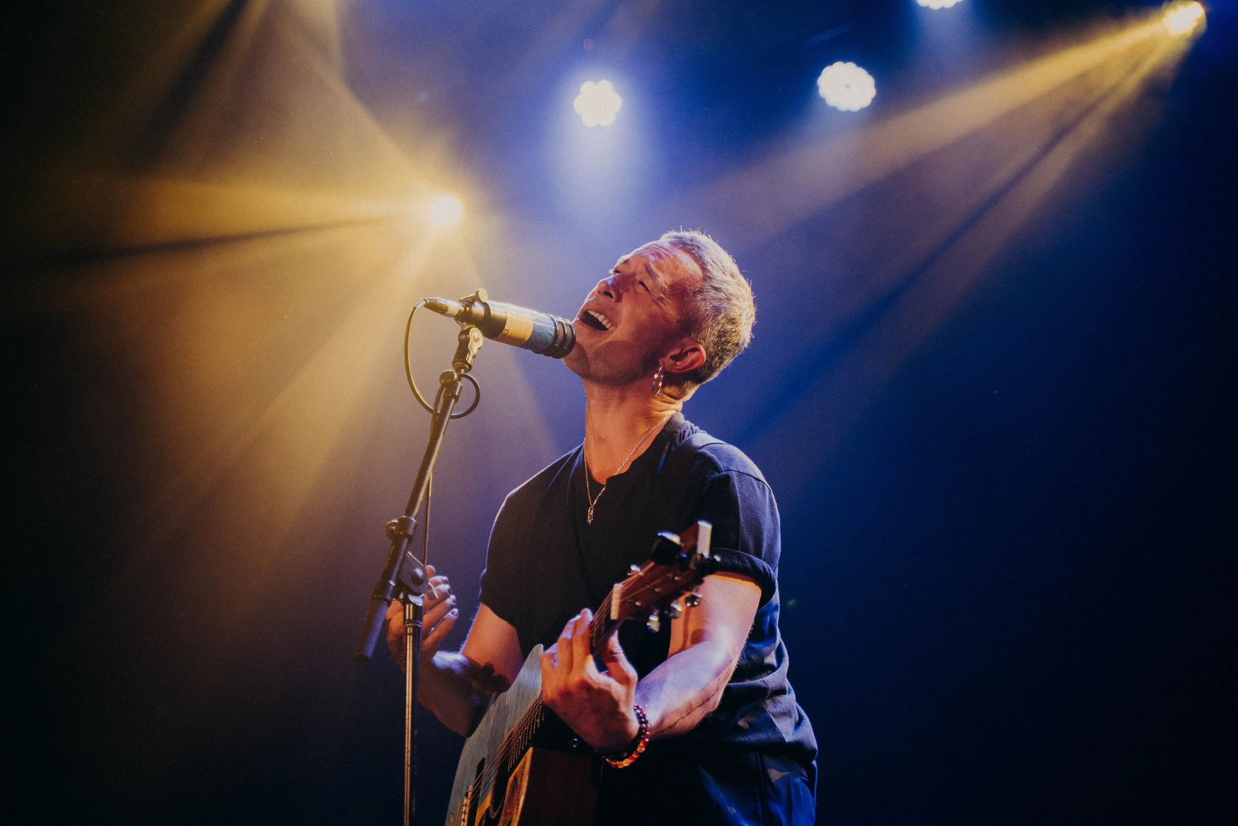 A male singer with short hair, earrings, a necklace, and a tattoo on his arm, passionately singing into a microphone while playing an acoustic guitar on stage, illuminated by yellow and blue stage lights.