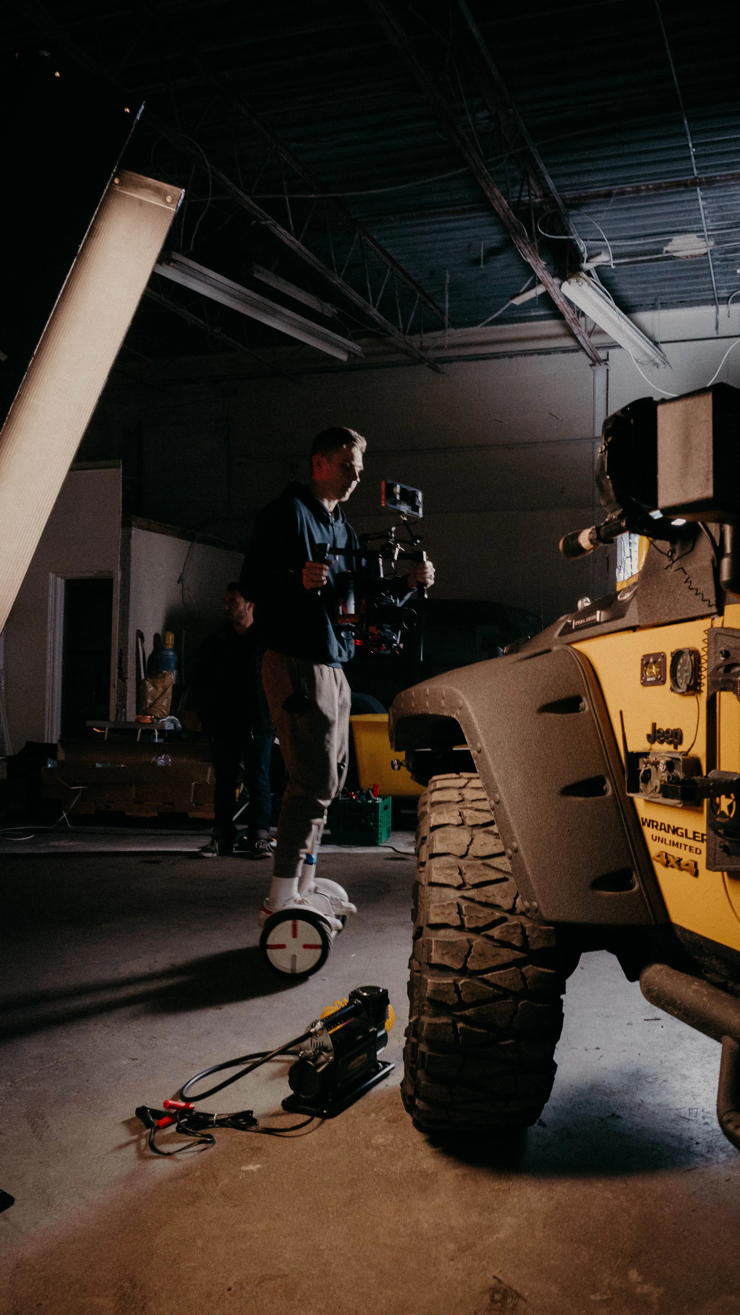 Person filming with a camera stabilizer in a warehouse-like setting with a yellow Jeep Wrangler in the foreground.