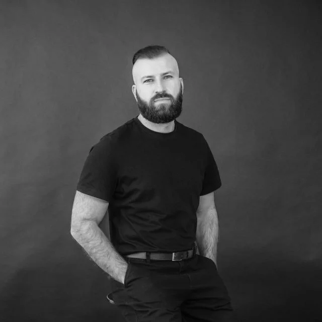 Black and white portrait of a man with a beard, wearing a black t-shirt and black pants, standing against a plain background