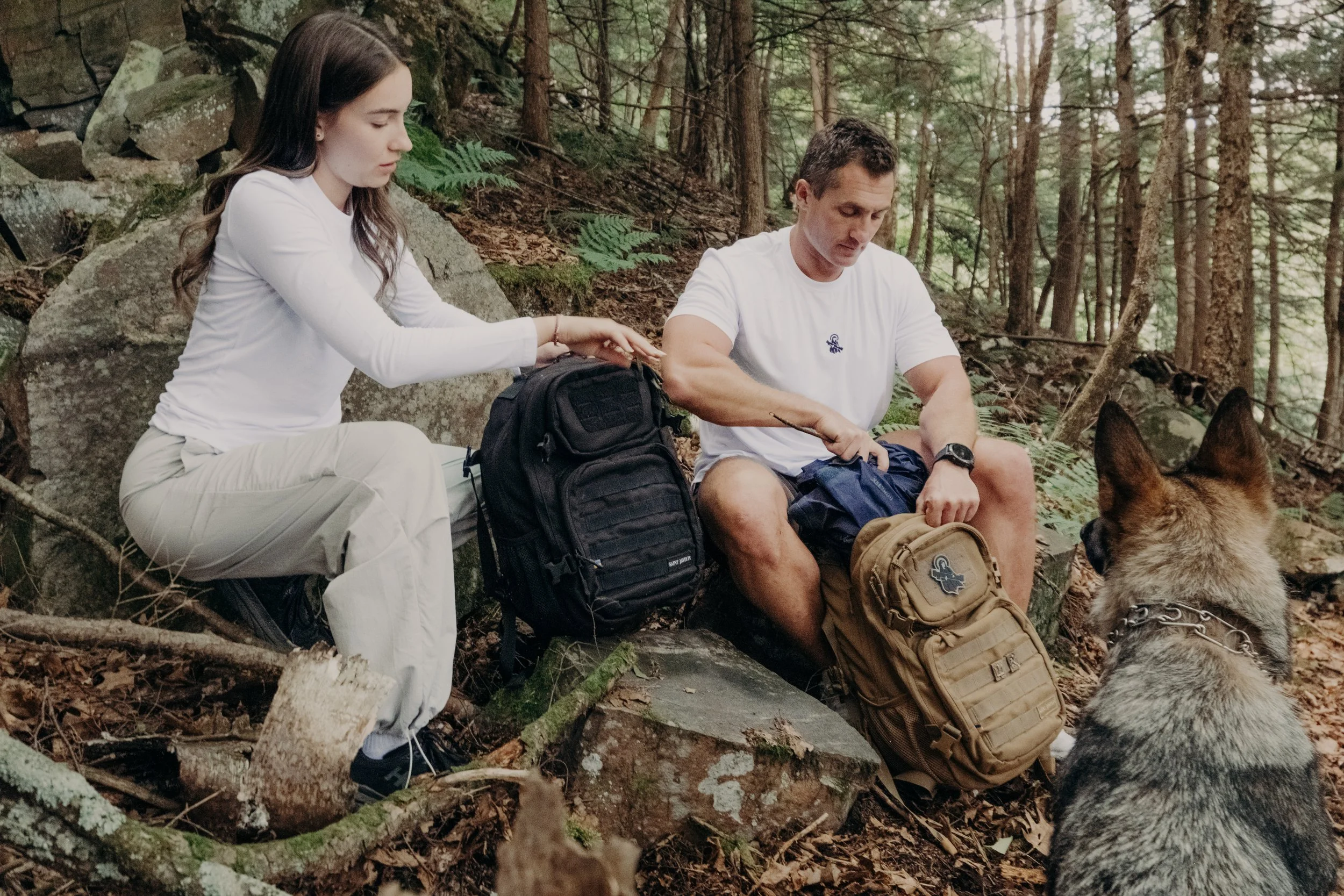 A man, woman, and a dog sitting outdoors in a wooded area, with the woman reaching into a backpack, all appearing to rest on rocks surrounded by trees and ferns.