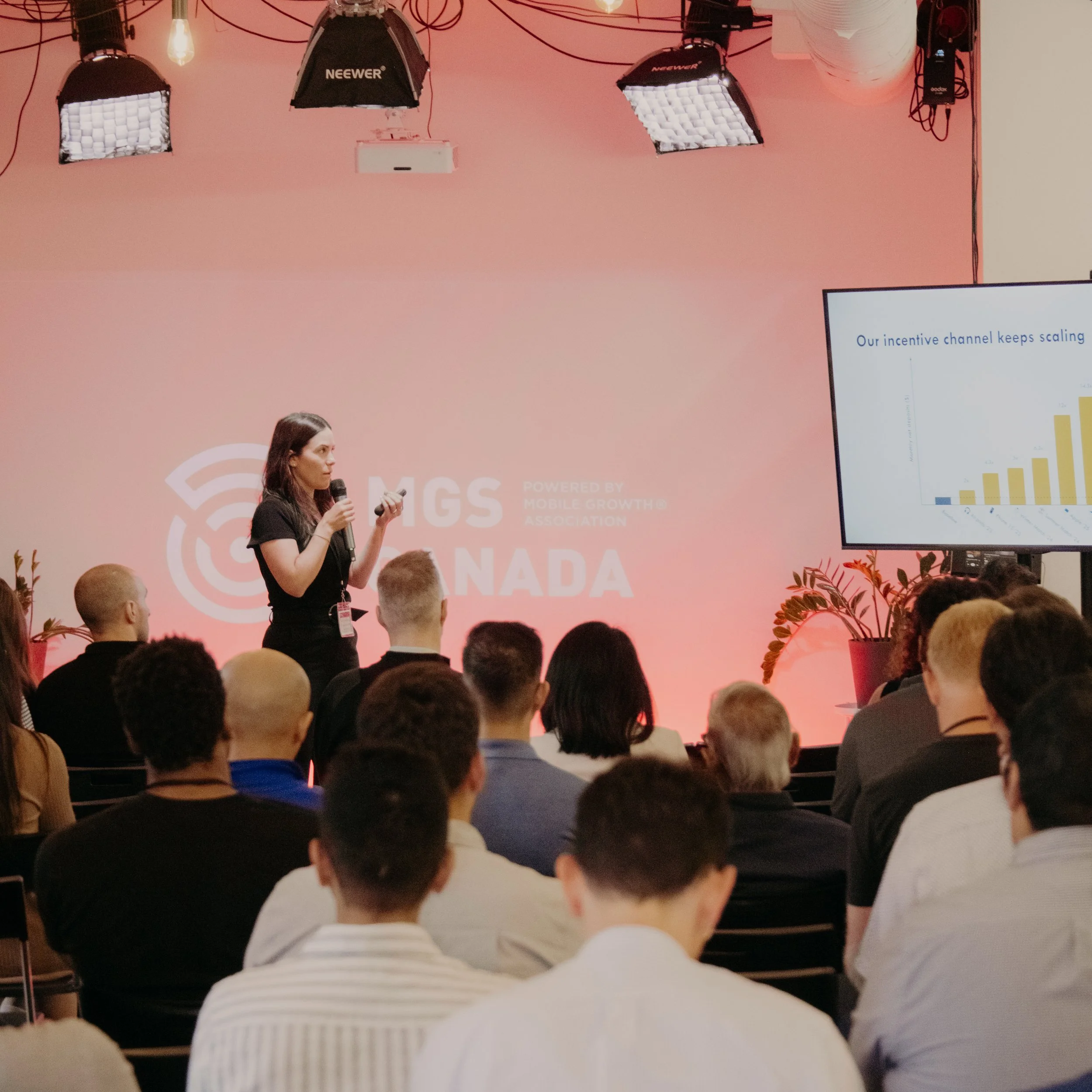 A woman giving a presentation to a group of people at a conference, standing in front of a pink backdrop with the logo 'MGS CANADA' and a screen displaying a bar graph.