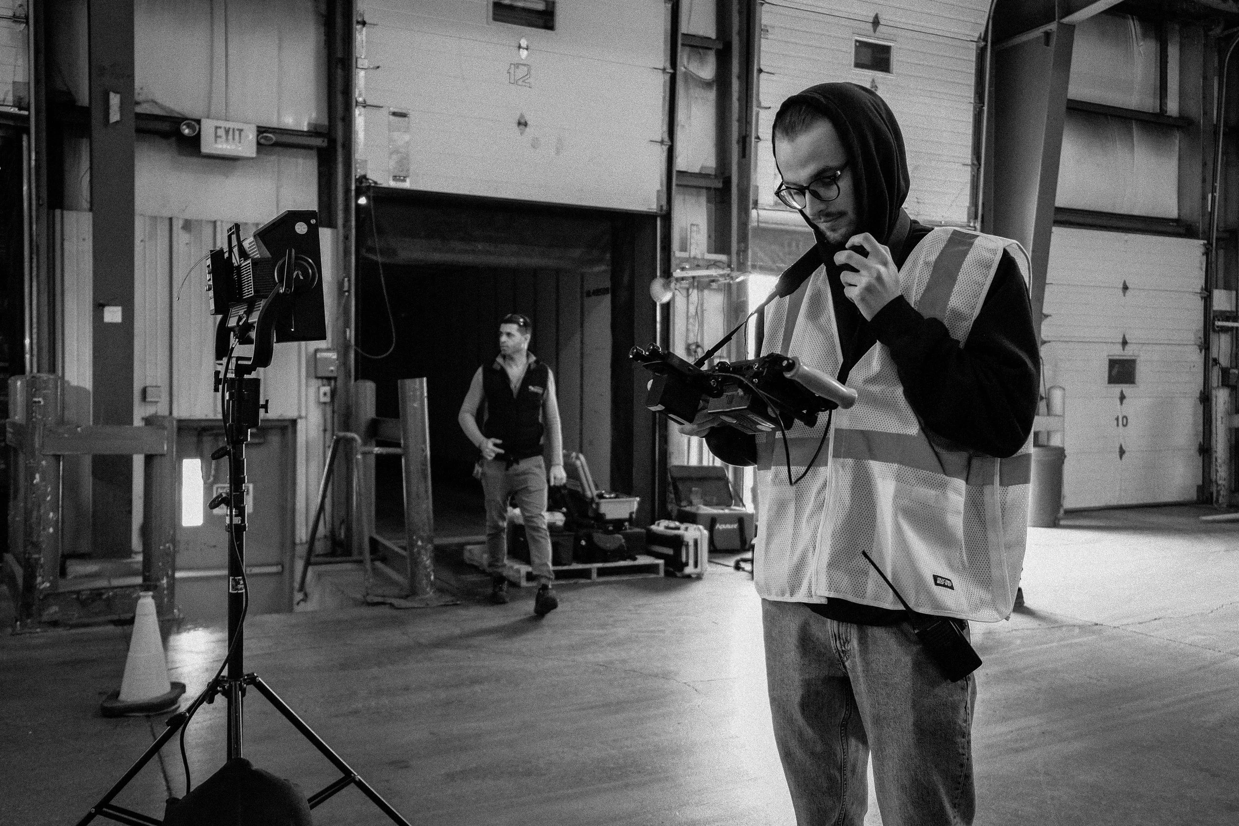 A man wearing glasses, a hoodie, and a reflective vest is operating a drone controller in an industrial warehouse. In the background, another person is standing near equipment, with large garage doors and a small cone nearby.