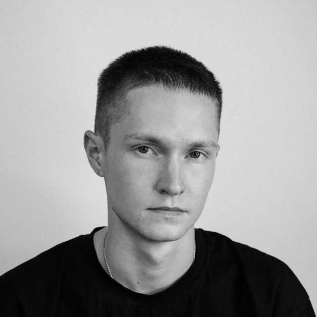Black and white portrait of a young man with short hair, wearing a black shirt and a chain necklace, looking directly at the camera.