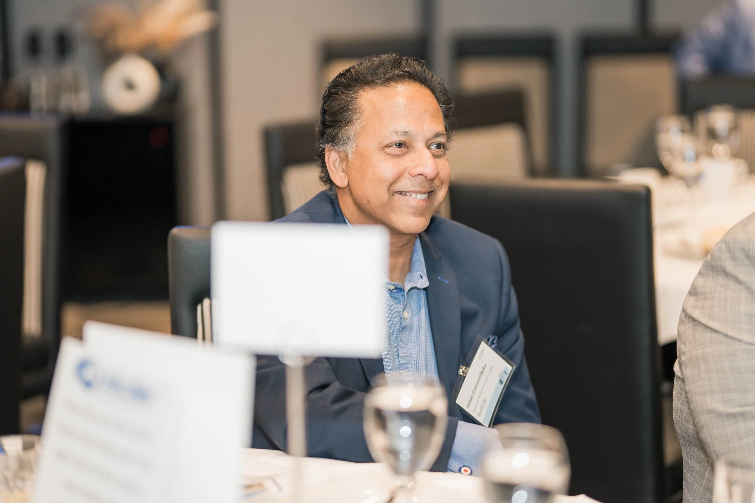 Man in a suit sitting at a conference table, smiling, with a name tag and glasses of water in front of him.
