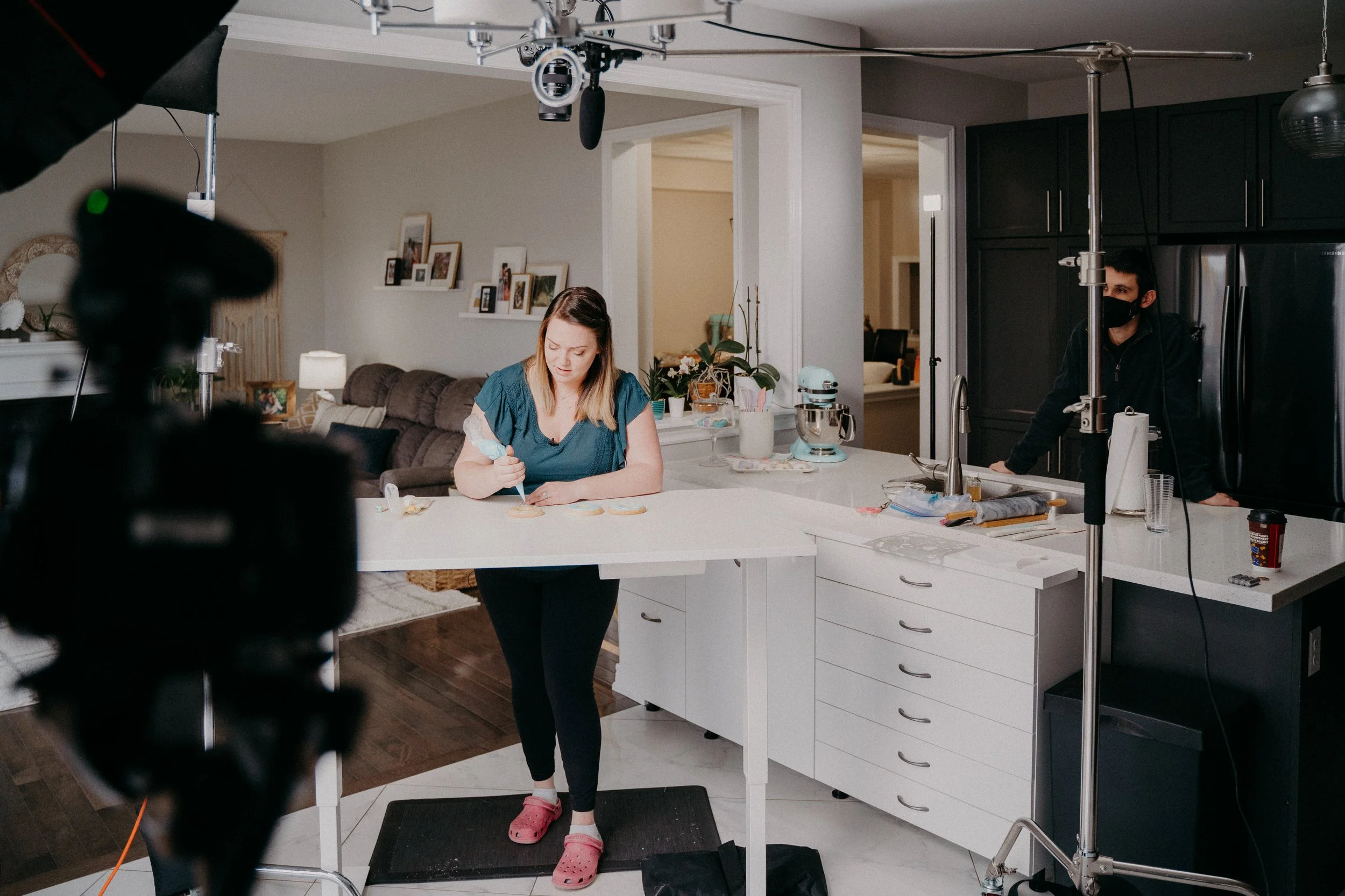 Filming of a cooking show in a modern kitchen. A woman is decorating cookies at a white kitchen island, while a man wearing a black mask stands near the fridge. Professional lighting and camera equipment are set up around them.