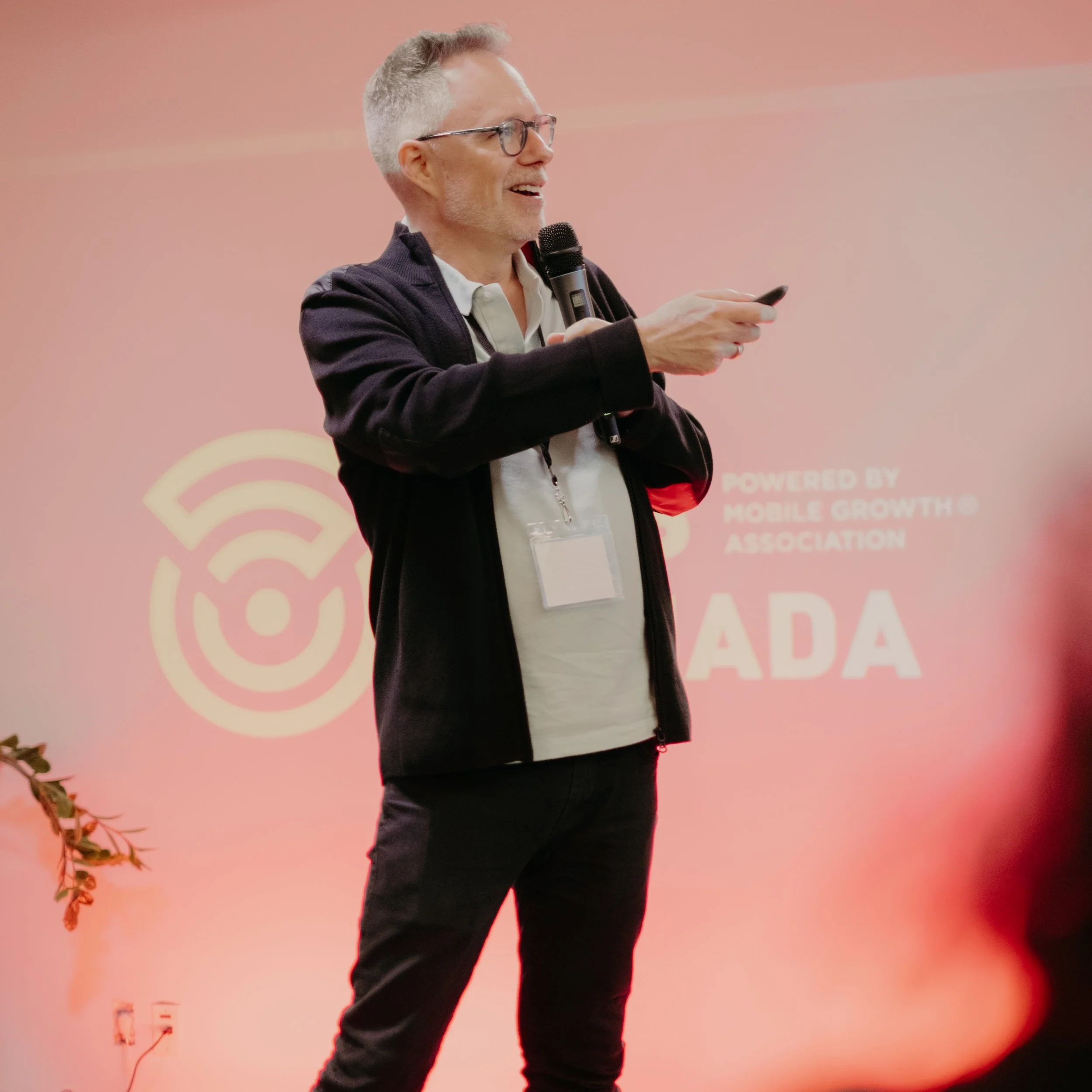 A man with glasses and gray hair speaking into a microphone and holding a phone, standing in front of a red backdrop with a logo and text.