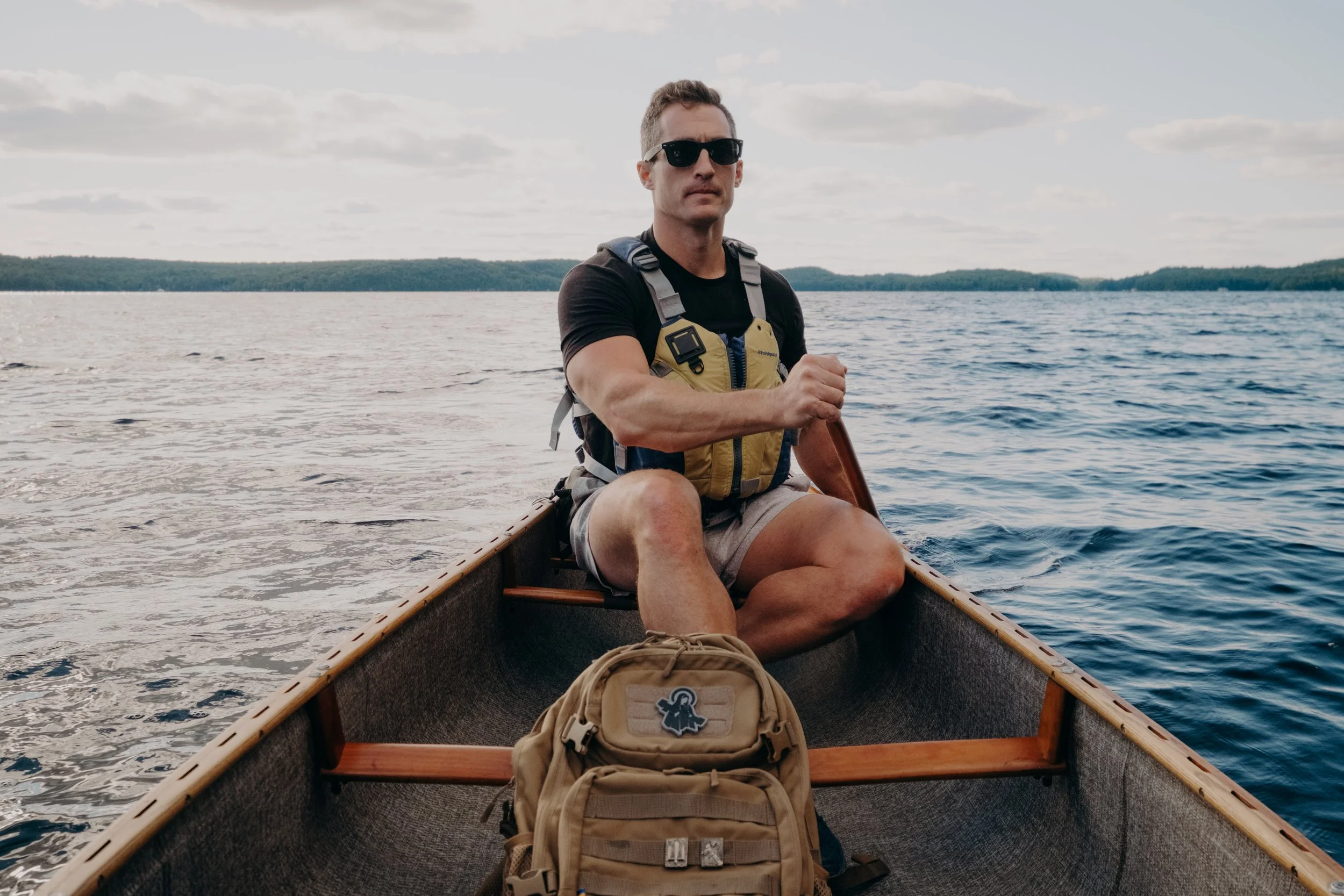 A man wearing sunglasses and a life jacket sits in a canoe on a lake, holding a paddle, with a backpack in the front of the canoe.