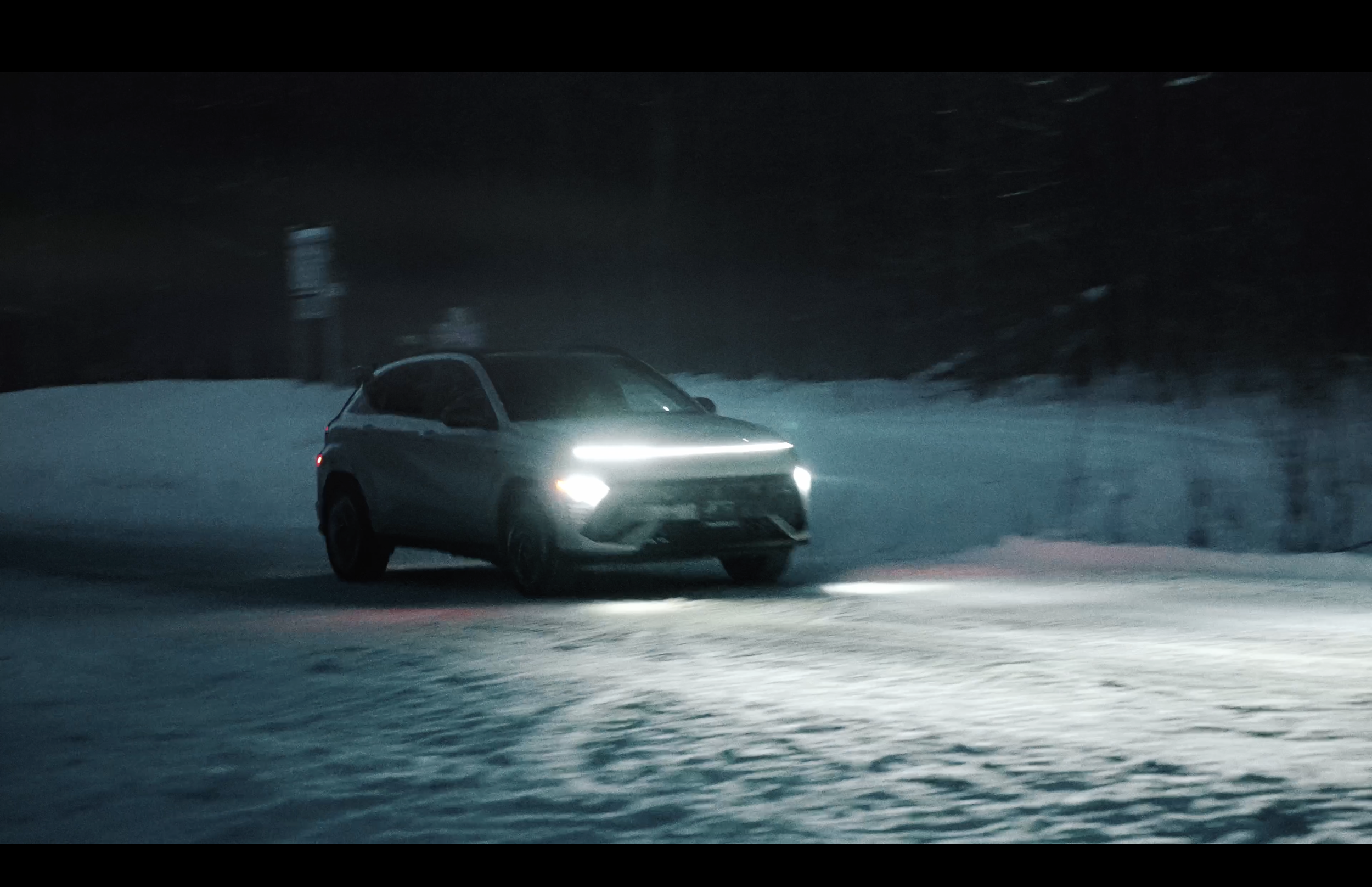 A silver SUV driving on a snow-covered road at night with its headlights on.