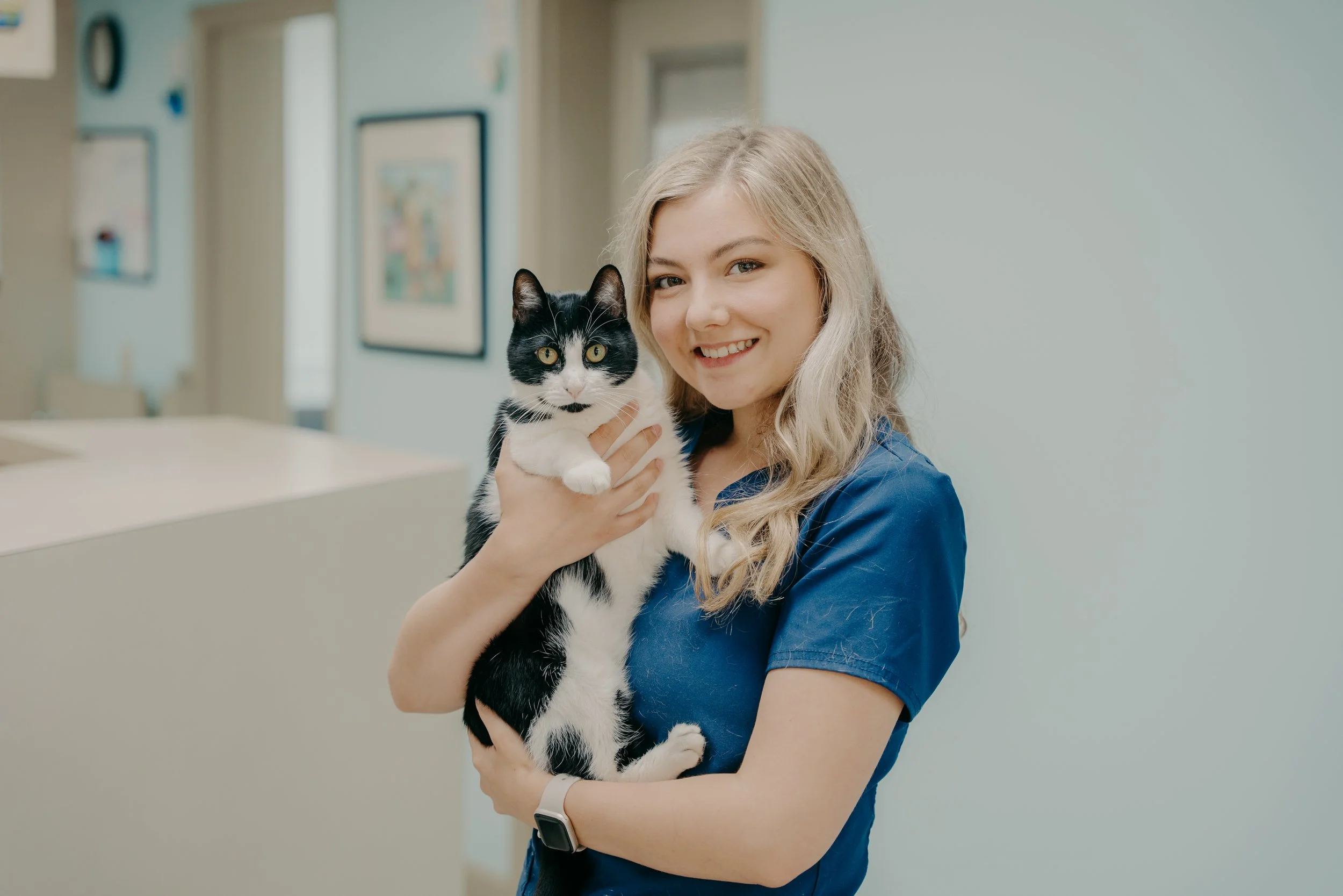 A woman in a blue uniform holding a black and white cat, smiling in a veterinary clinic or animal care setting.