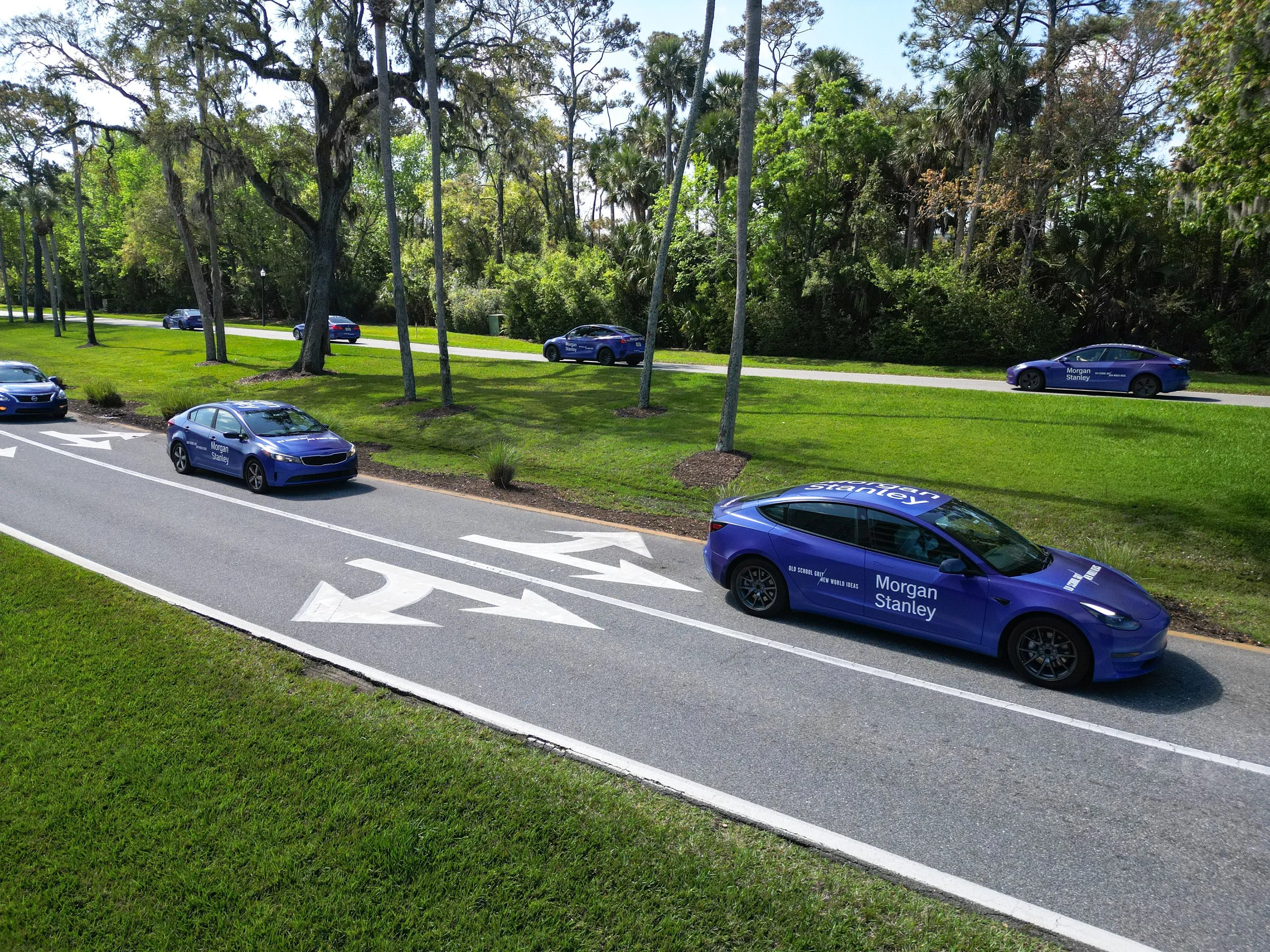 Multiple blue cars with Morgan Stanley branding on a multi-lane road with green grass, trees, and a wooded area in the background.