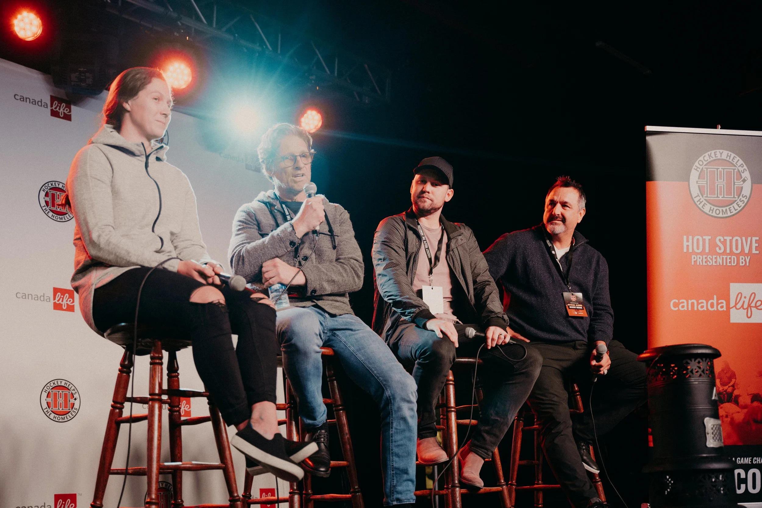 Four people sitting on barstools on stage during a panel discussion, with microphones in hand. The backdrop features the logos for Canada Life and Hockey Helps the Homeless, and a banner reads 'Hot Stove Presented by Canada Life'.