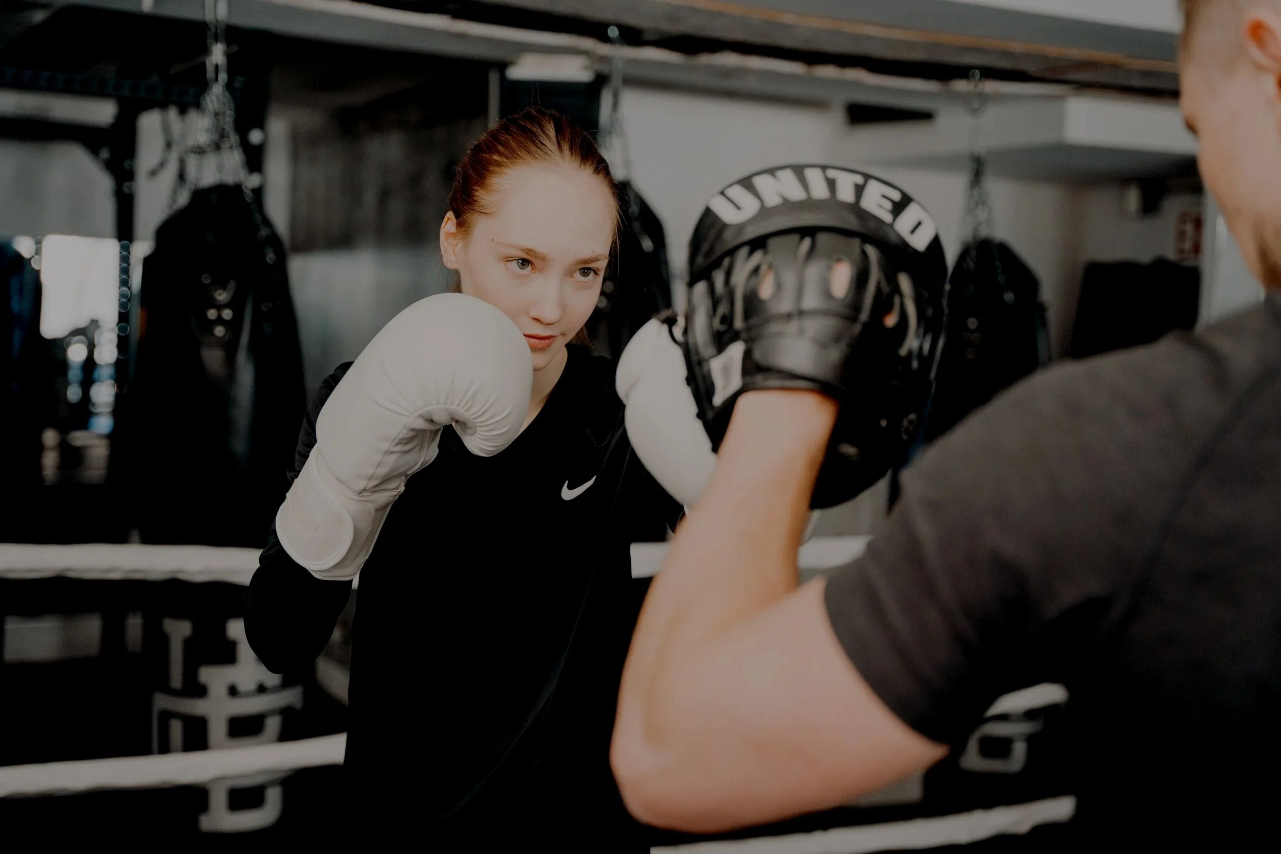 A young woman with red hair and wearing boxing gloves and athletic clothing practicing boxing with a trainer wearing a black helmet and gloves in a gym.