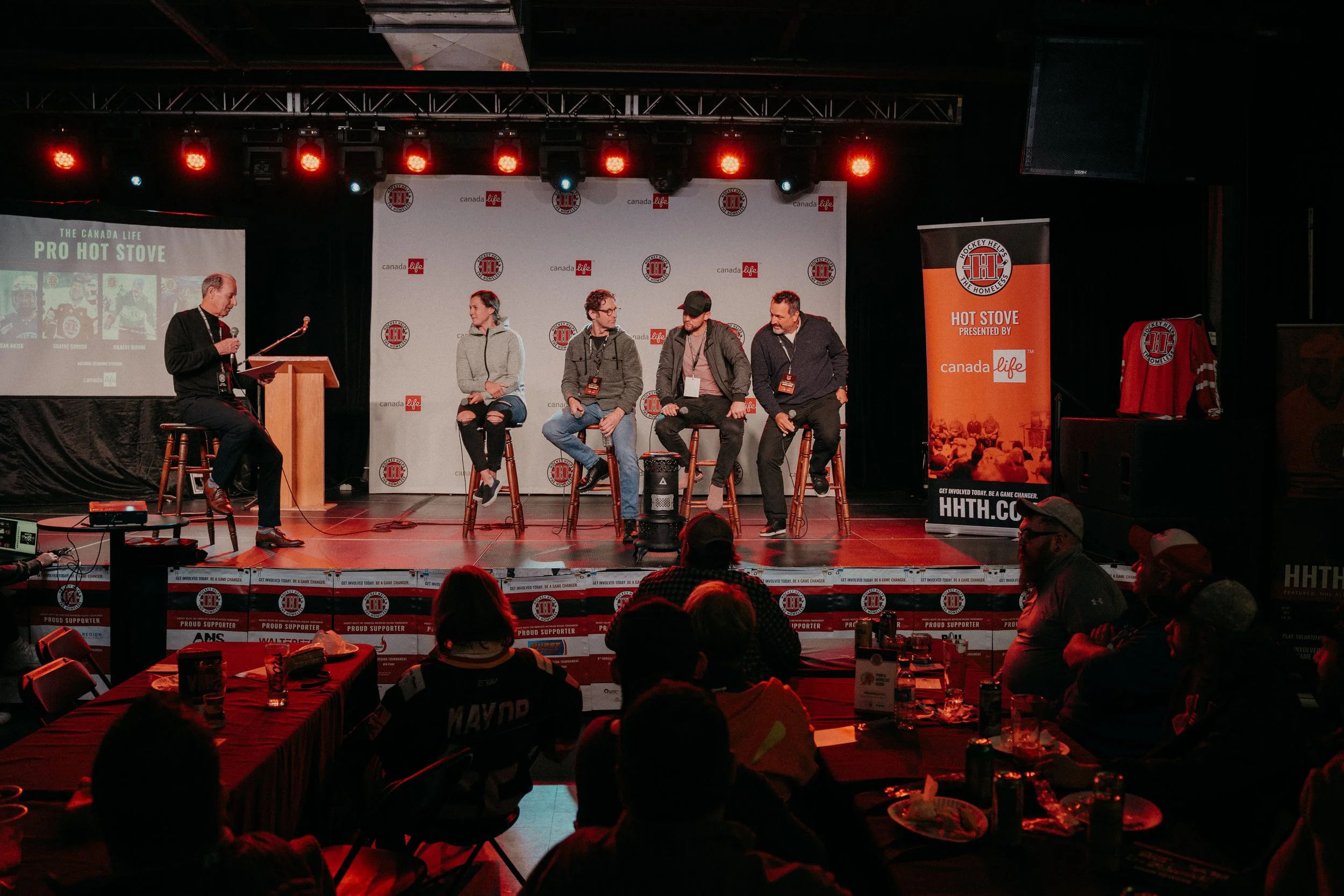 Panel discussion on stage with five people seated on stools, a moderator on a separate stool with a microphone, audience seated in front, and banners and screens related to Canada's hot stove hockey event.