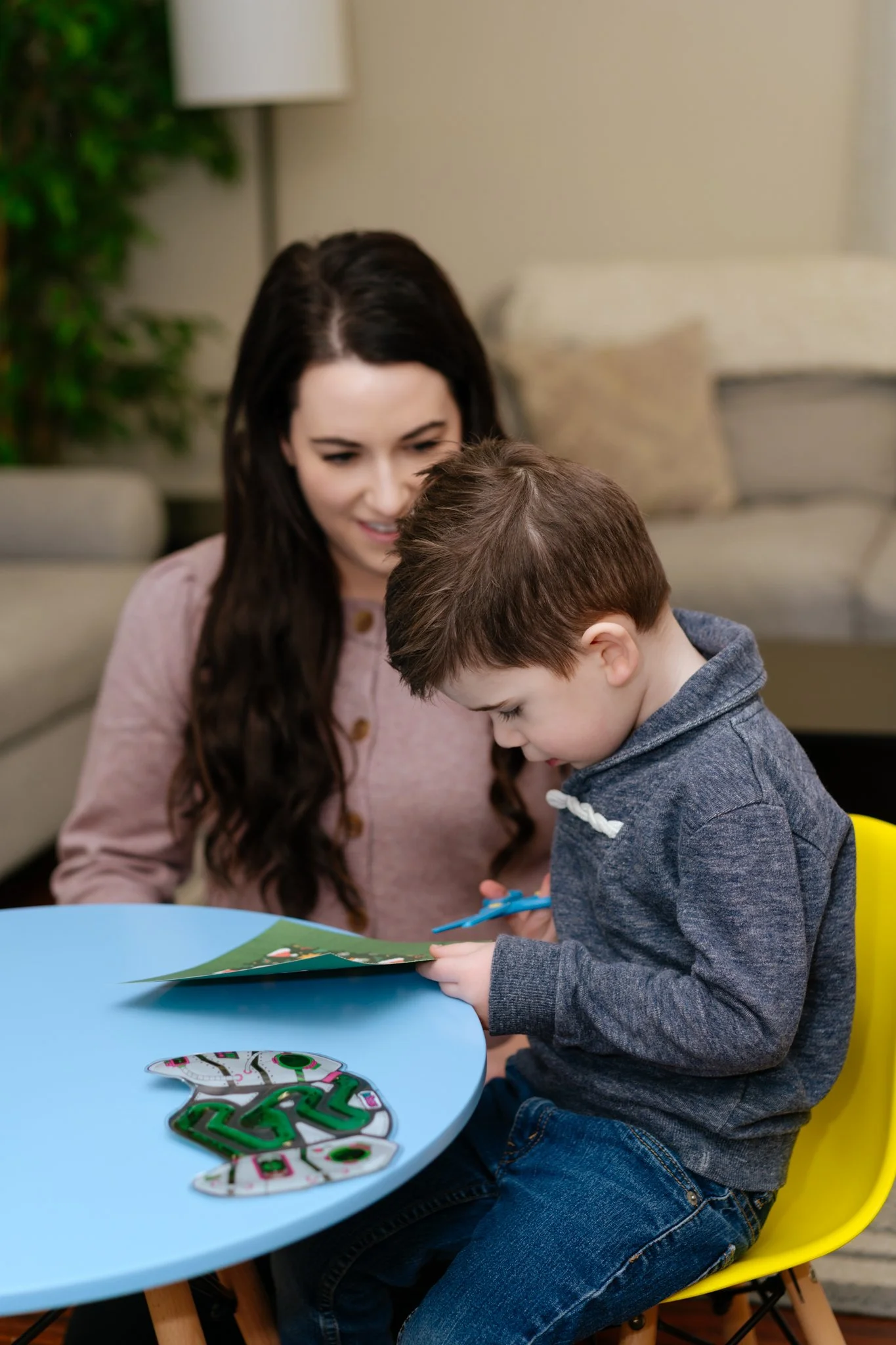 A woman and a young boy seated at a small blue table. The boy is cutting a Christmas-themed card with scissors, and there are some Christmas tree puzzle pieces on the table.