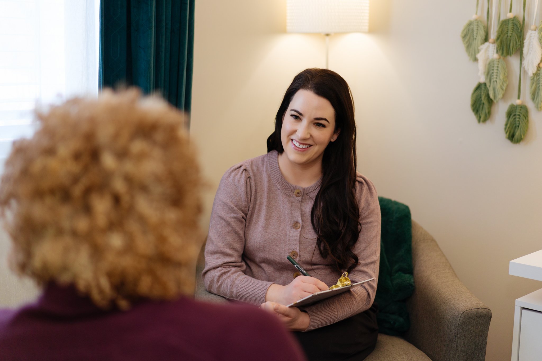 A woman with long dark hair sitting on a beige couch, smiling and taking notes on a clipboard during a consultation, with another person with curly blond hair partially visible in the foreground.