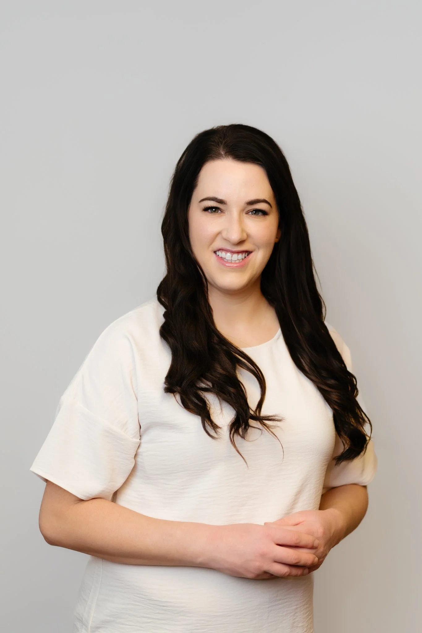 A woman with long dark hair, wearing a light-colored top, smiling and standing against a gray background.
