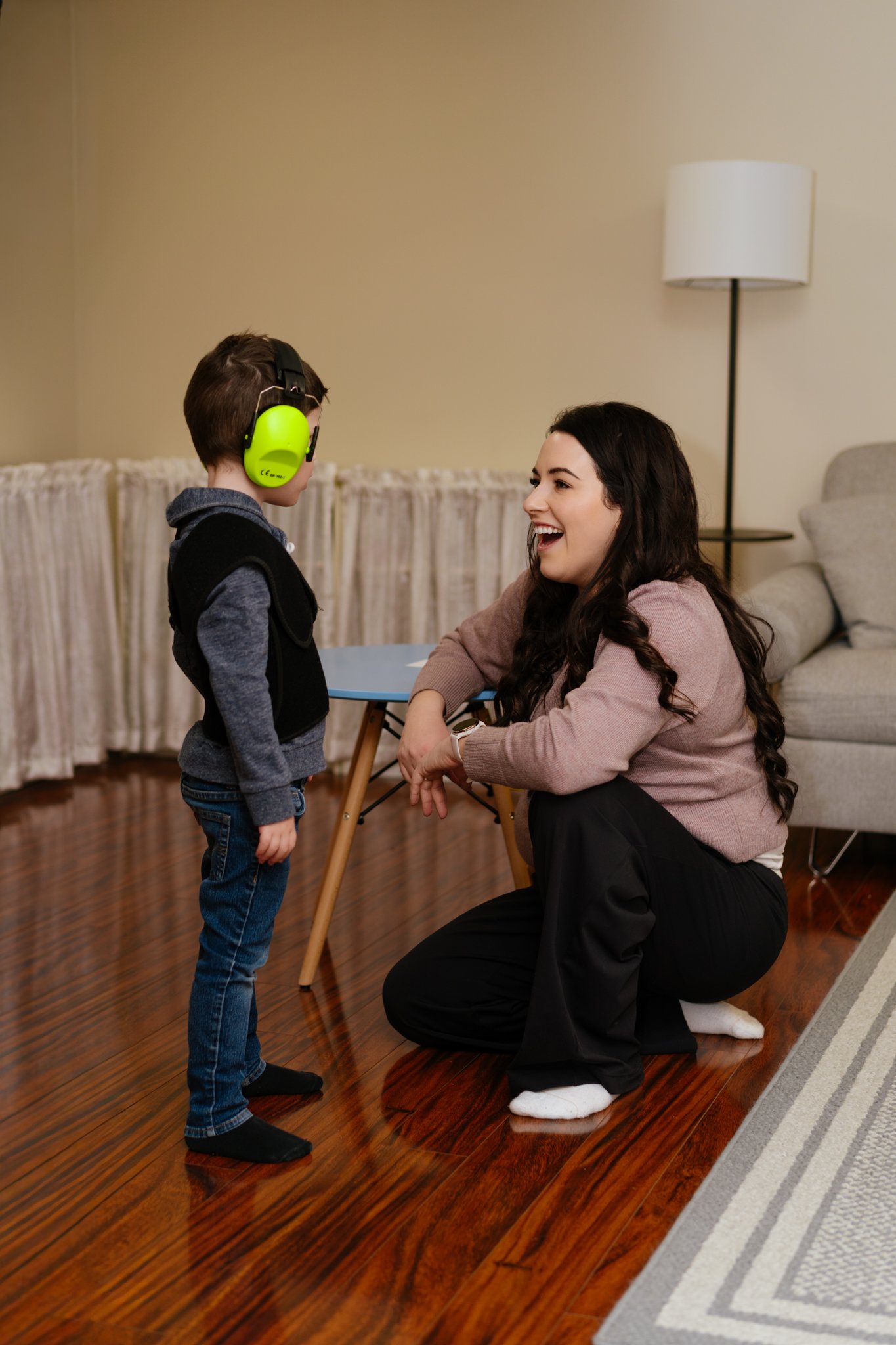A woman and a young boy sharing a joyful moment indoors, with the boy wearing bright green noise-canceling headphones.