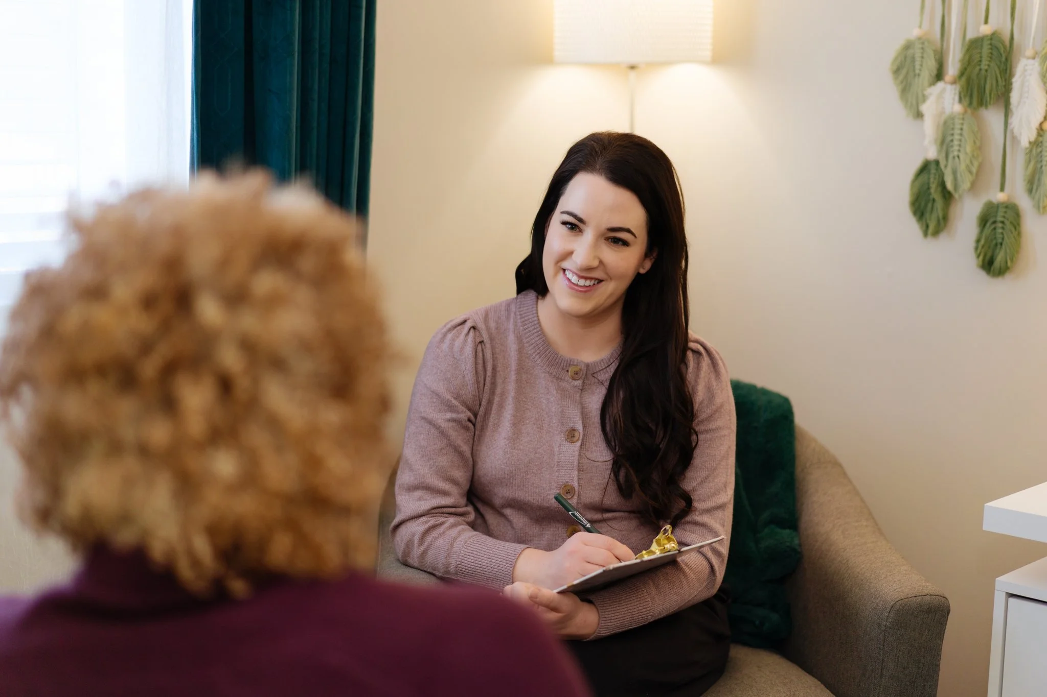 A woman with long dark hair smiling and taking notes during a consultation with an older woman with curly blonde hair in a cozy, well-lit room.